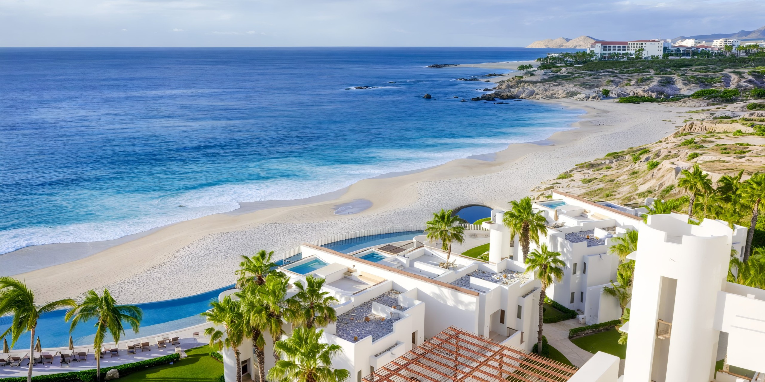 Aerial view of the Marquis Los Cabos resort, featuring white buildings, palm trees, and an infinity pool overlooking a sandy beach and blue ocean under a clear sky.