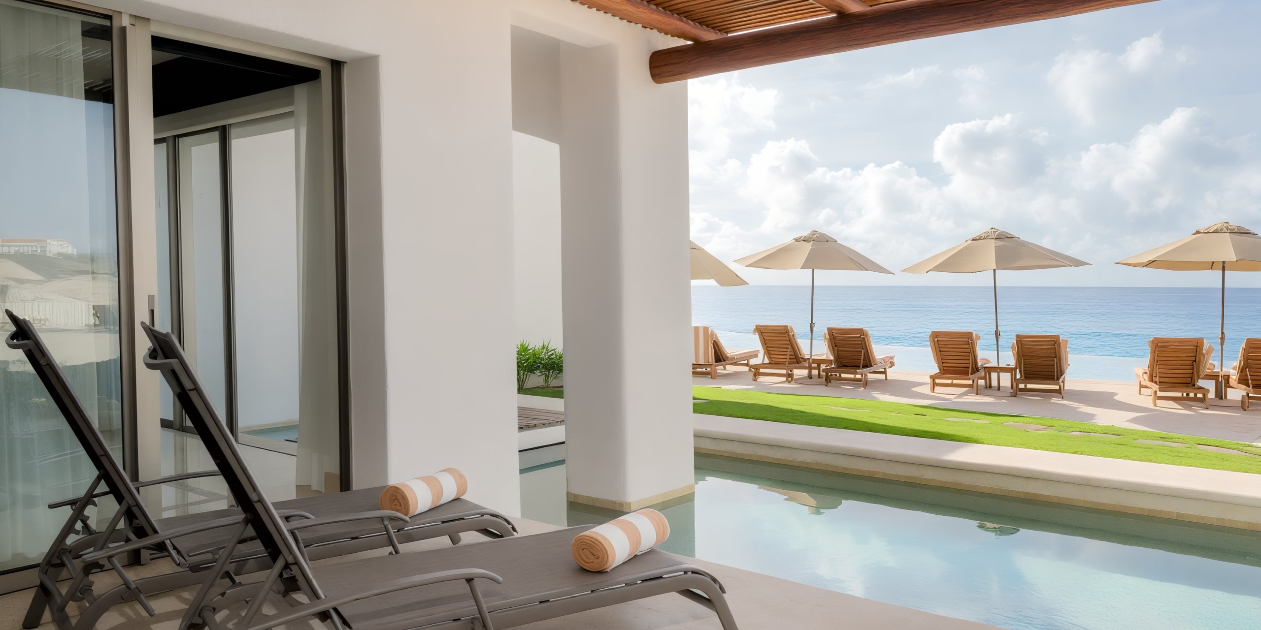 Experience luxury at Marquis Los Cabos, featuring an elegant outdoor pool area with lounge chairs and umbrellas overlooking a serene sea, all adjacent to a minimalist white building and wooden pergola.