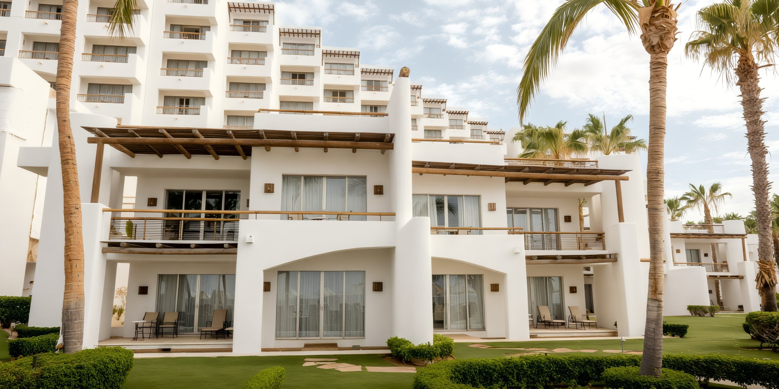 The Marquis Los Cabos is a white multi-story hotel featuring balconies and lush palm trees at its entrance, surrounded by manicured lawns under a partly cloudy sky.