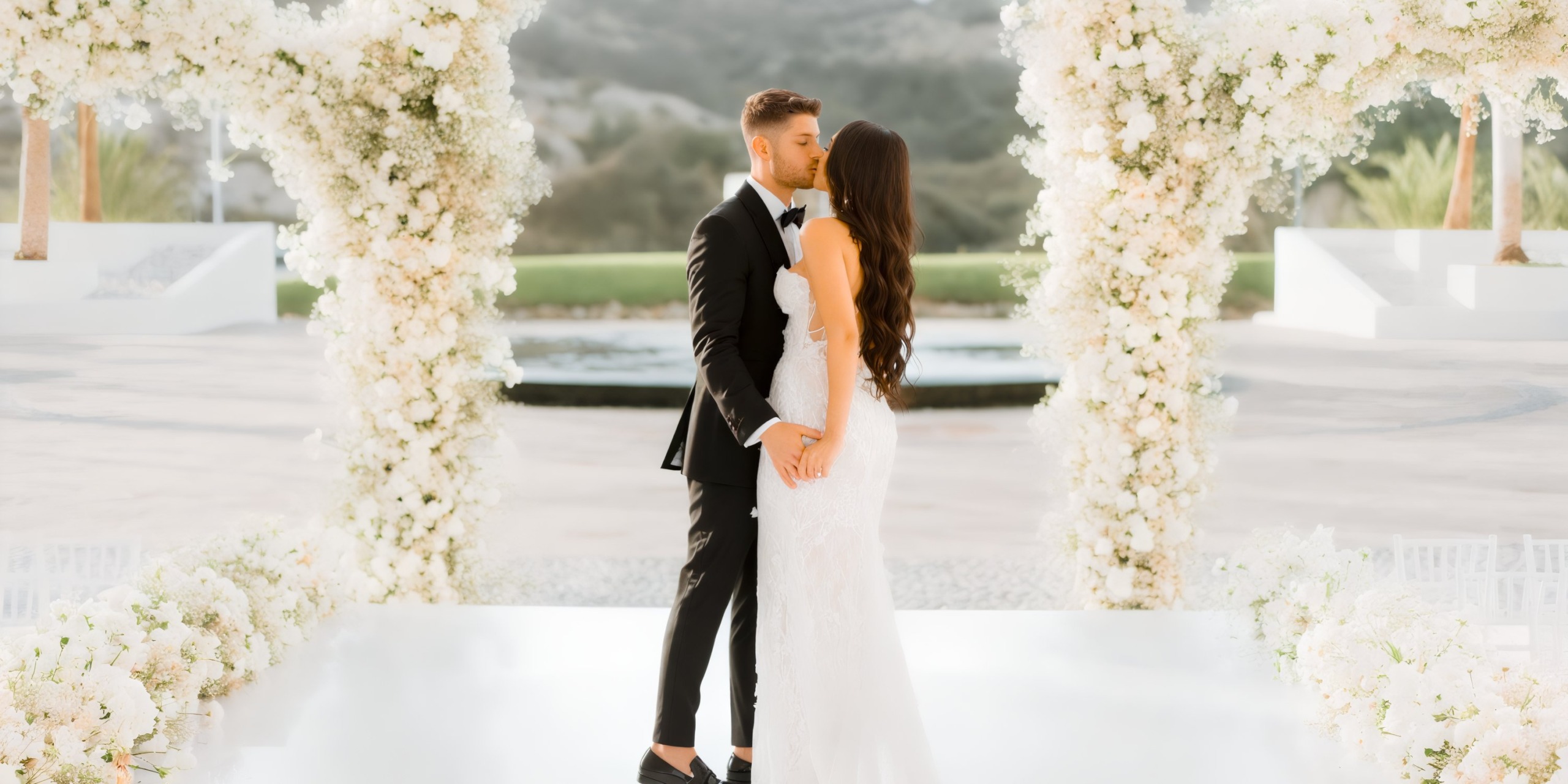 A couple in wedding attire shares a kiss under a floral arch on an outdoor platform at Marquis Los Cabos, with a blurred natural background.