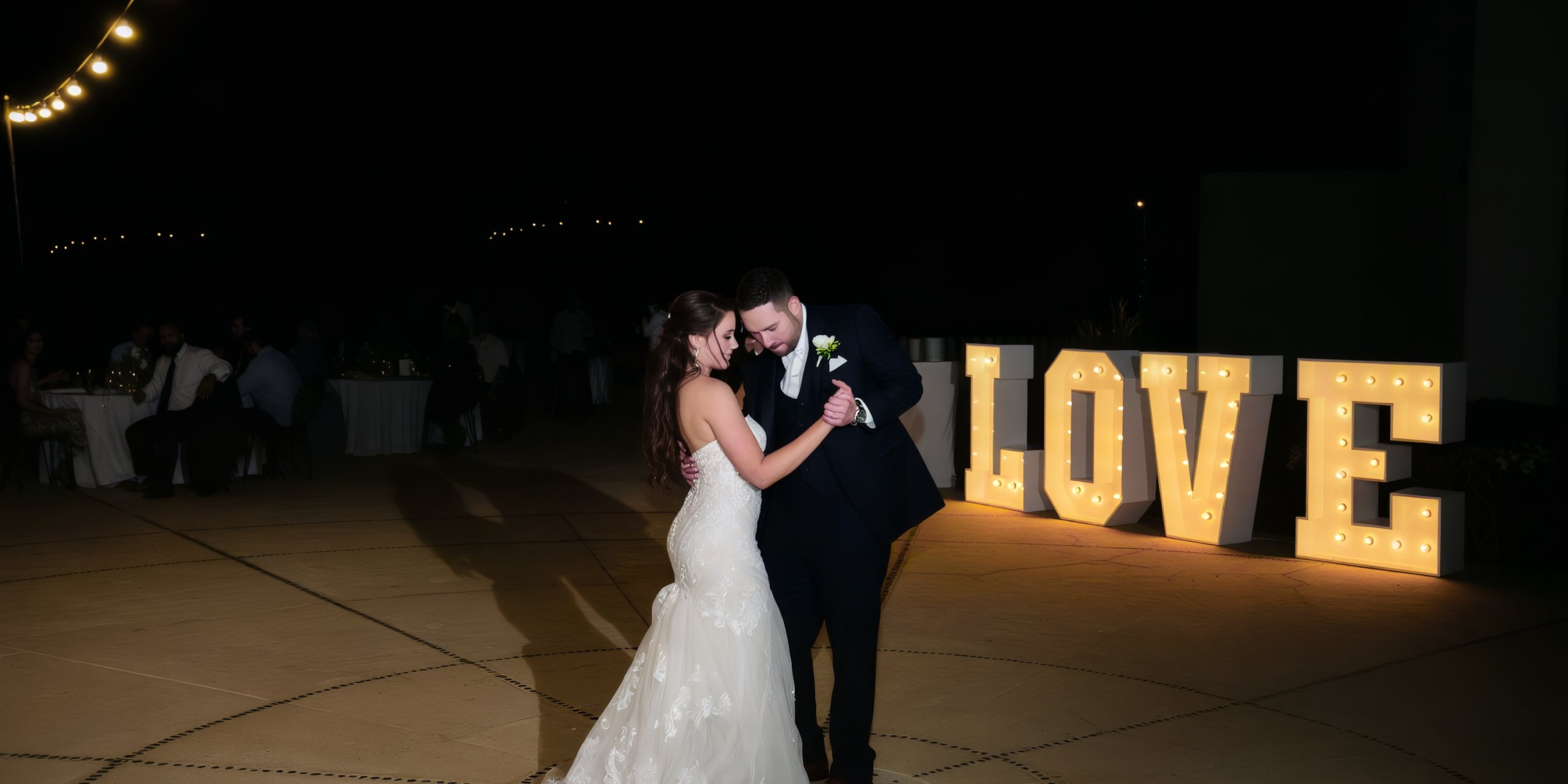 A couple in formal attire dances outdoors at night near large illuminated letters spelling "LOVE" at Marquis Los Cabos.