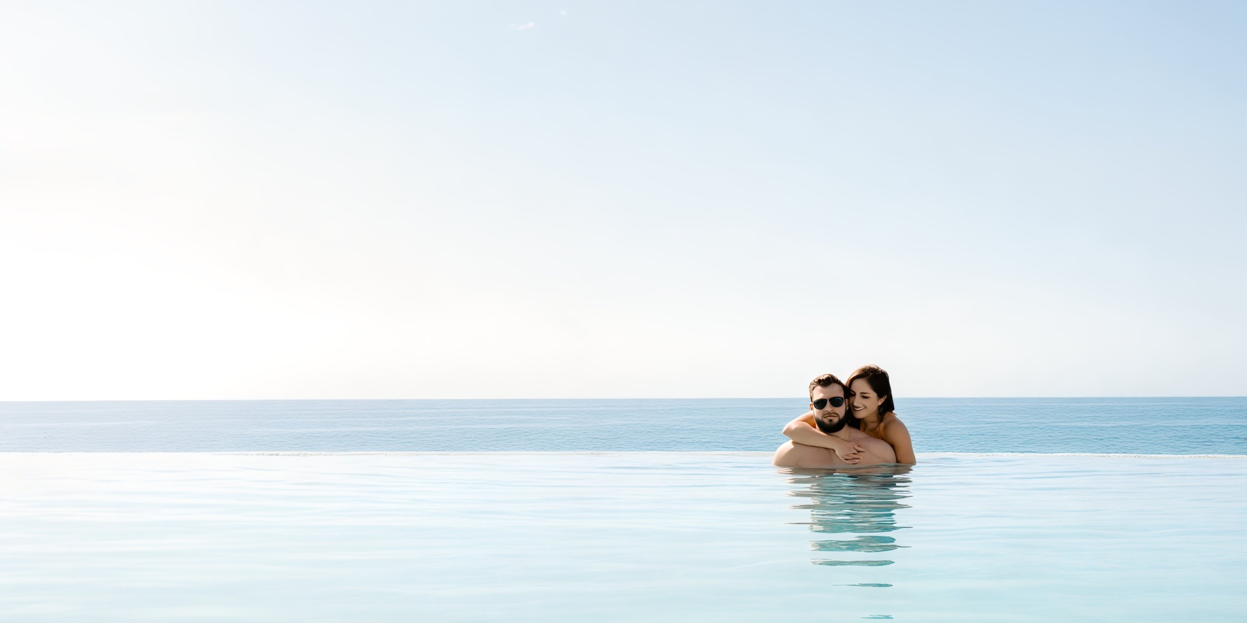 A couple embraces in the infinity pool at Marquis Los Cabos, enjoying a stunning ocean view under a clear sky.