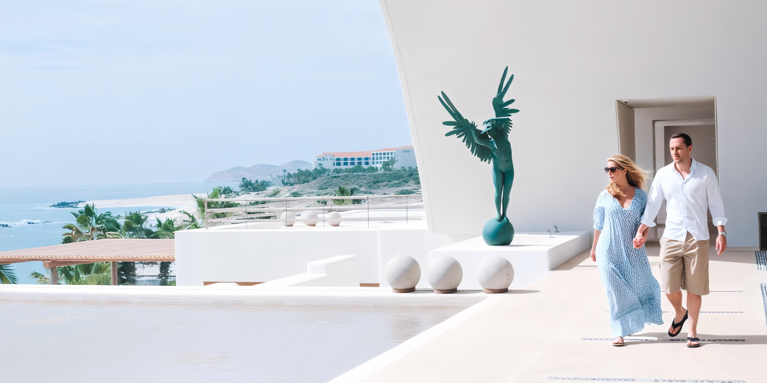 A couple strolls along the modern terrace of Marquis Los Cabos by the ocean, passing a winged statue.