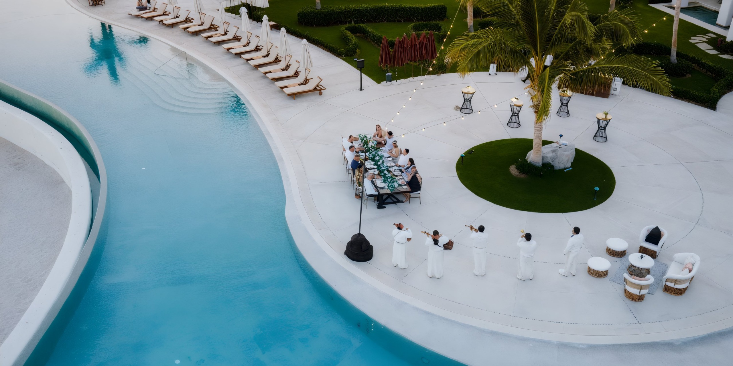 An aerial view at Marquis Los Cabos reveals a group dining at a long table beside a large pool, surrounded by empty loungers. Musicians in white perform under string lights, while a palm tree and spherical sculptures decorate the scene.