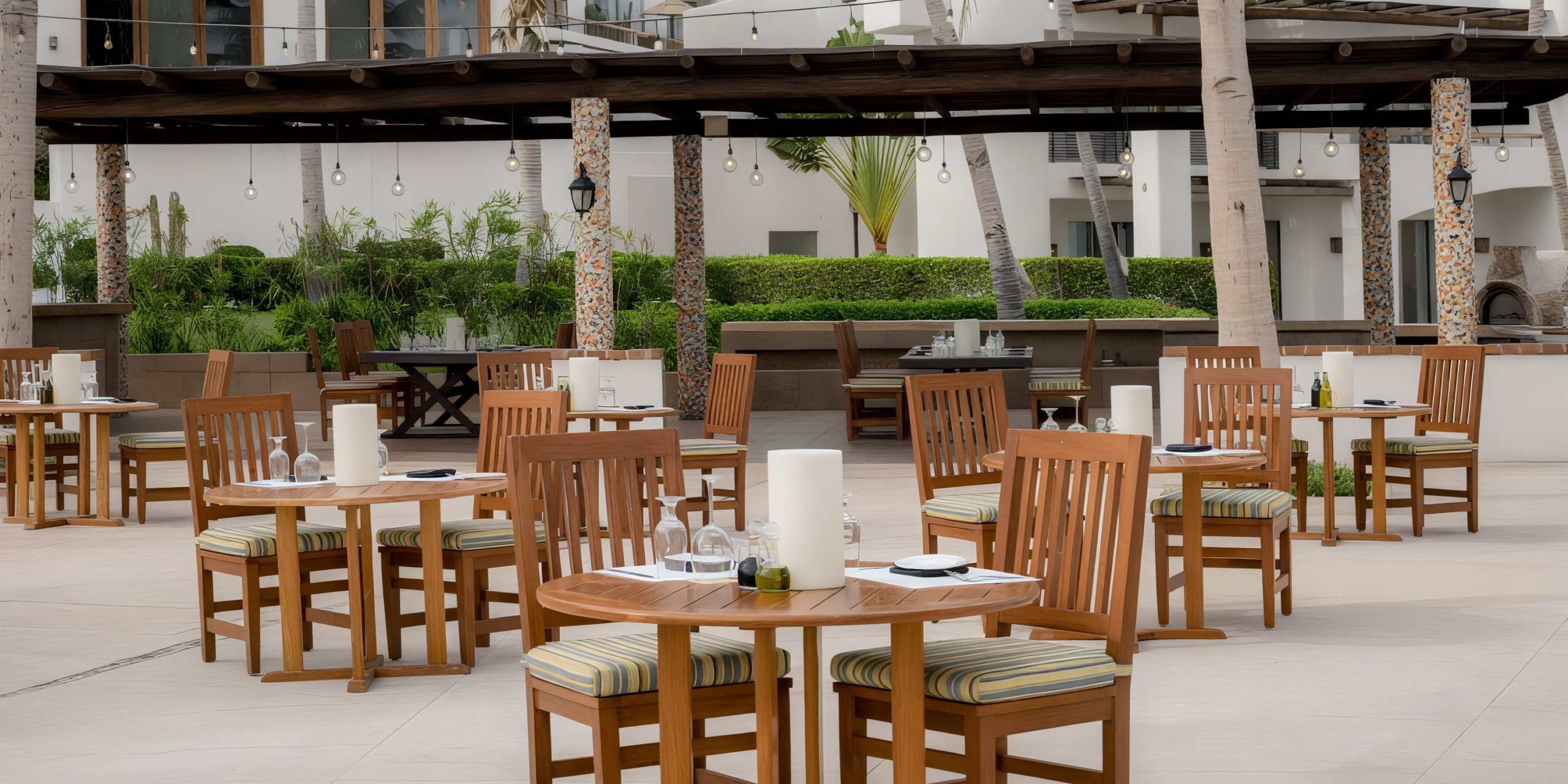 The outdoor restaurant patio at Marquis Los Cabos features wooden tables and chairs. Tables are set with glasses, plates, and centerpieces, surrounded by greenery and complemented by a building with a shaded area in the background.