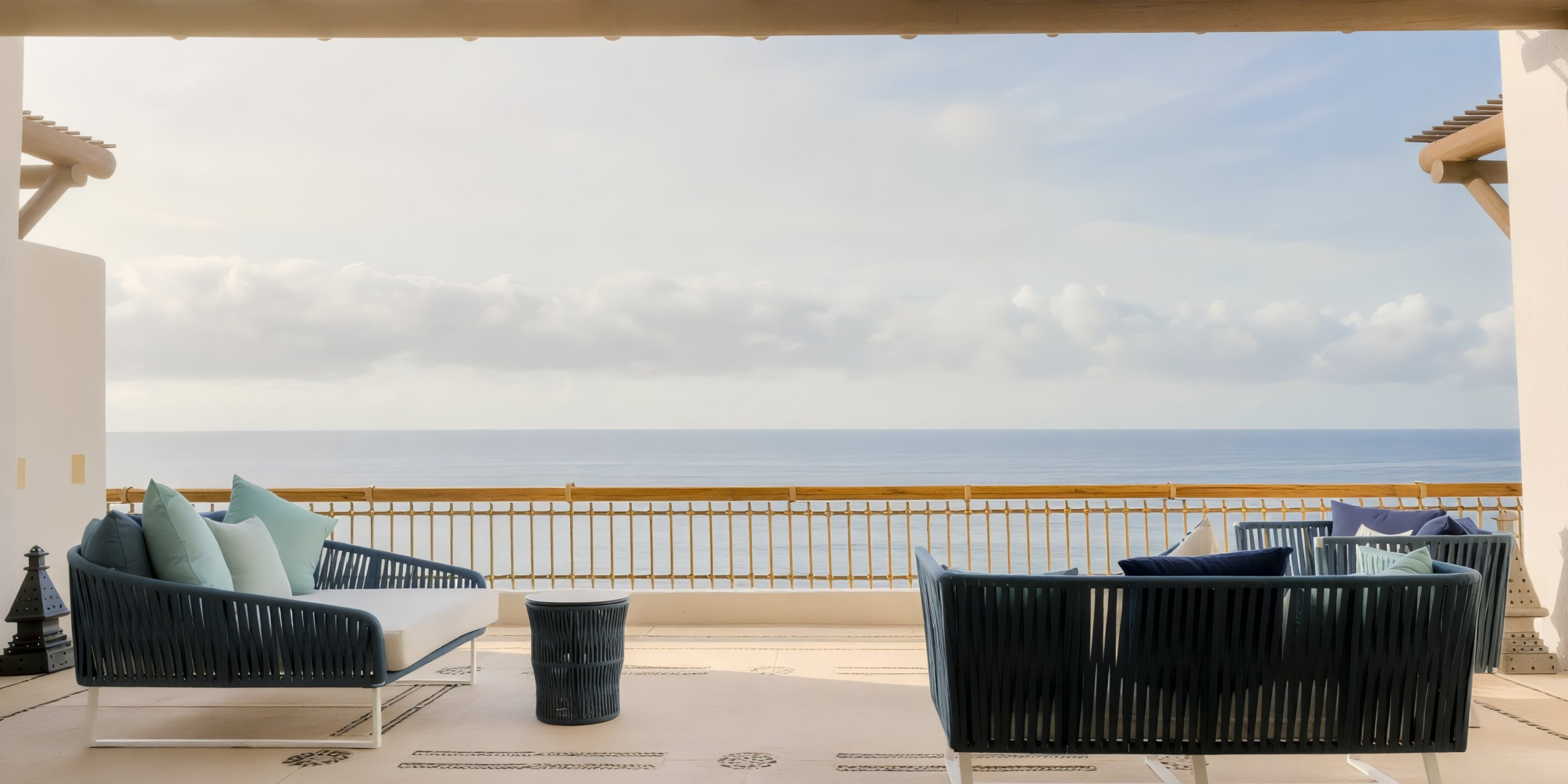 On the Marquis Los Cabos' outdoor patio, you'll find wicker furniture, a small table, and decorative pillows, all overlooking a serene ocean under a cloudy sky.