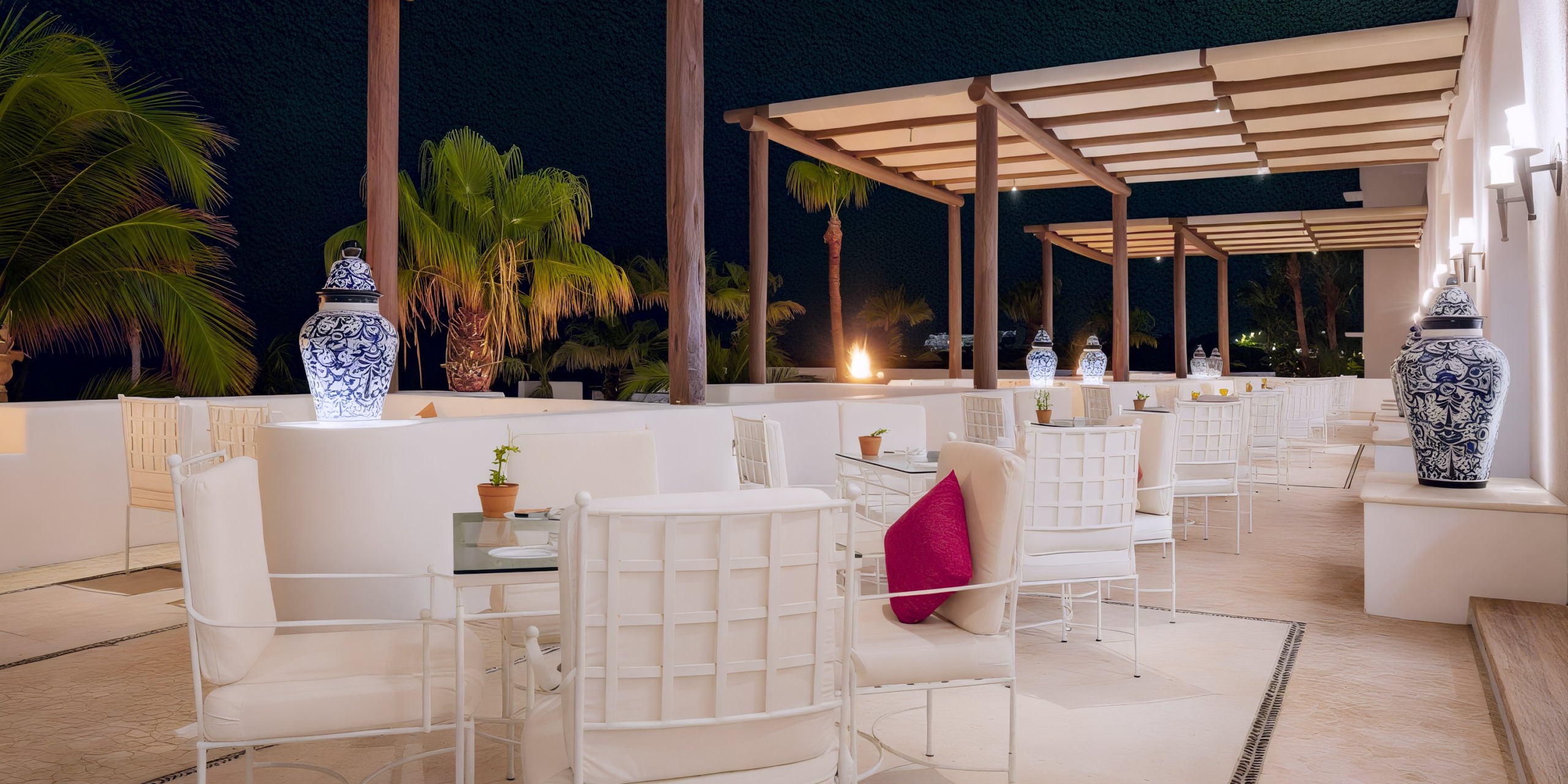 An outdoor area at Marquis Los Cabos features white chairs and tables, a pink cushion, decorative vases, palm trees, and a pergola.