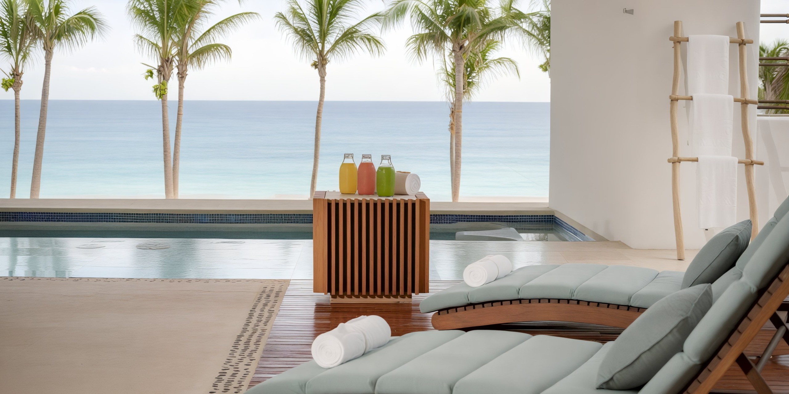 At Marquis Los Cabos, enjoy a poolside view with two lounge chairs and a wooden table adorned with juice bottles and rolled towels, set against a backdrop of palm trees and the ocean.