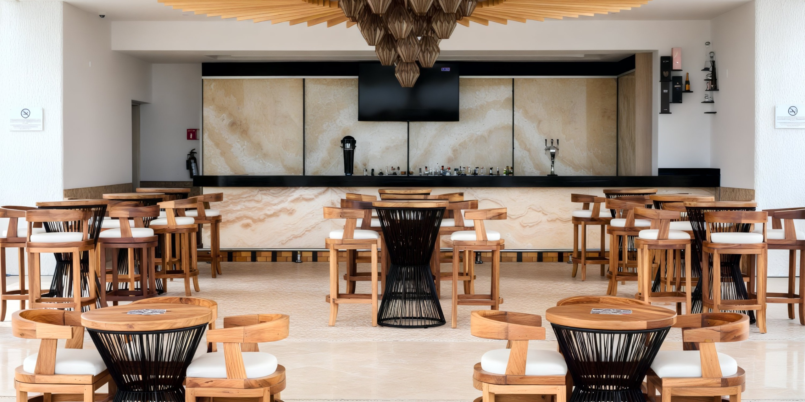 Modern bar area inspired by Marquis Los Cabos, featuring light wood furniture, a central black wire chair, decorative ceiling elements, and a bar counter with a TV screen and drink dispensers.