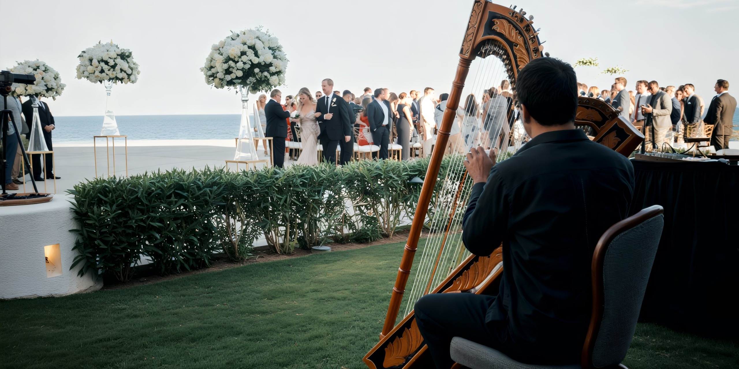 At an outdoor wedding reception at Marquis Los Cabos near the ocean, a harpist plays while guests gather around a table adorned with white floral decorations.
