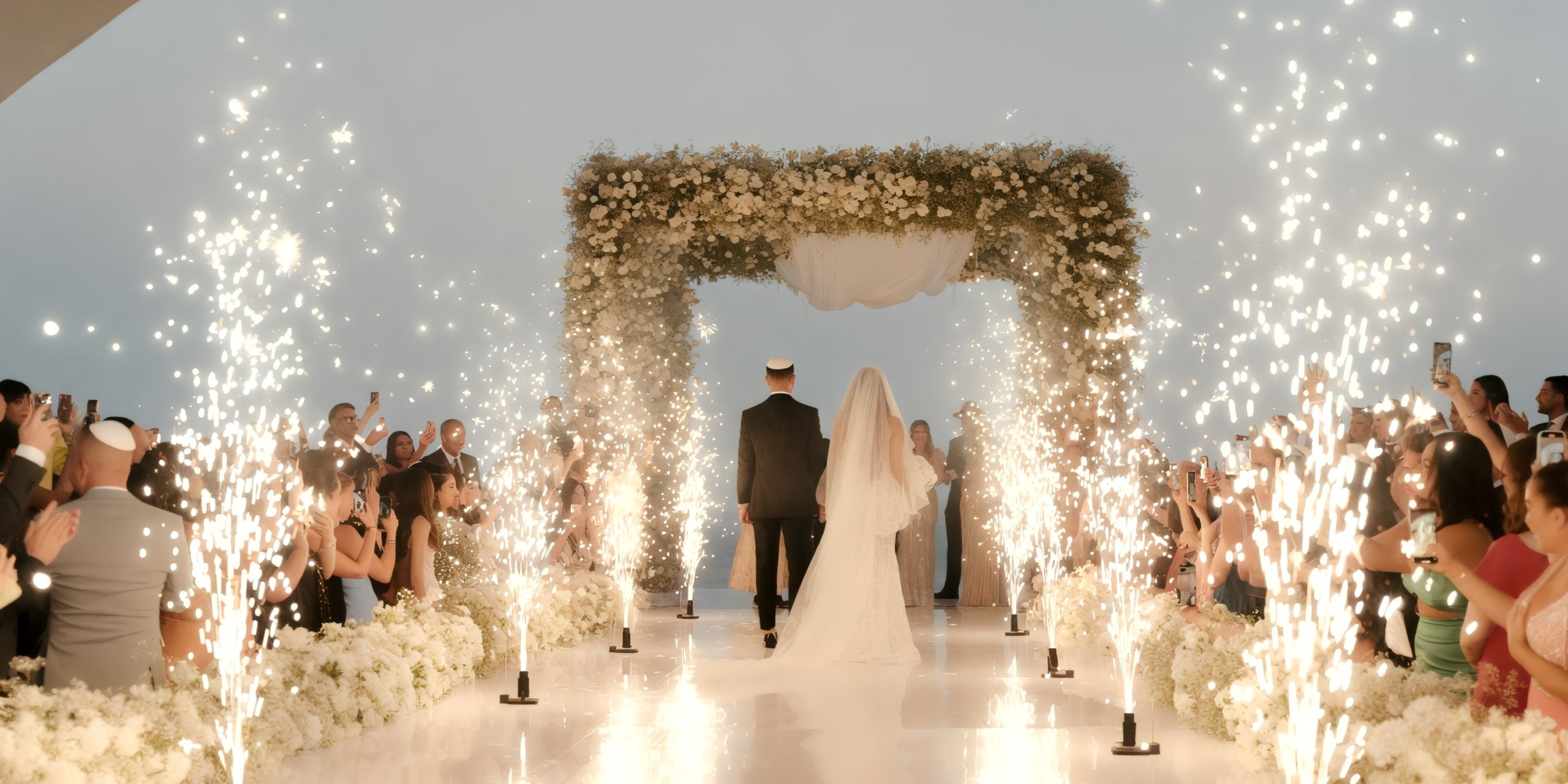 The bride and groom stroll down the aisle at Marquis Los Cabos beneath a flower-adorned archway, surrounded by guests and dazzling fireworks.