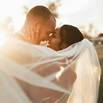 The radiant bride and groom share an embrace, their veil elegantly flowing in the sunlight, capturing the enchanting essence of a destination wedding.