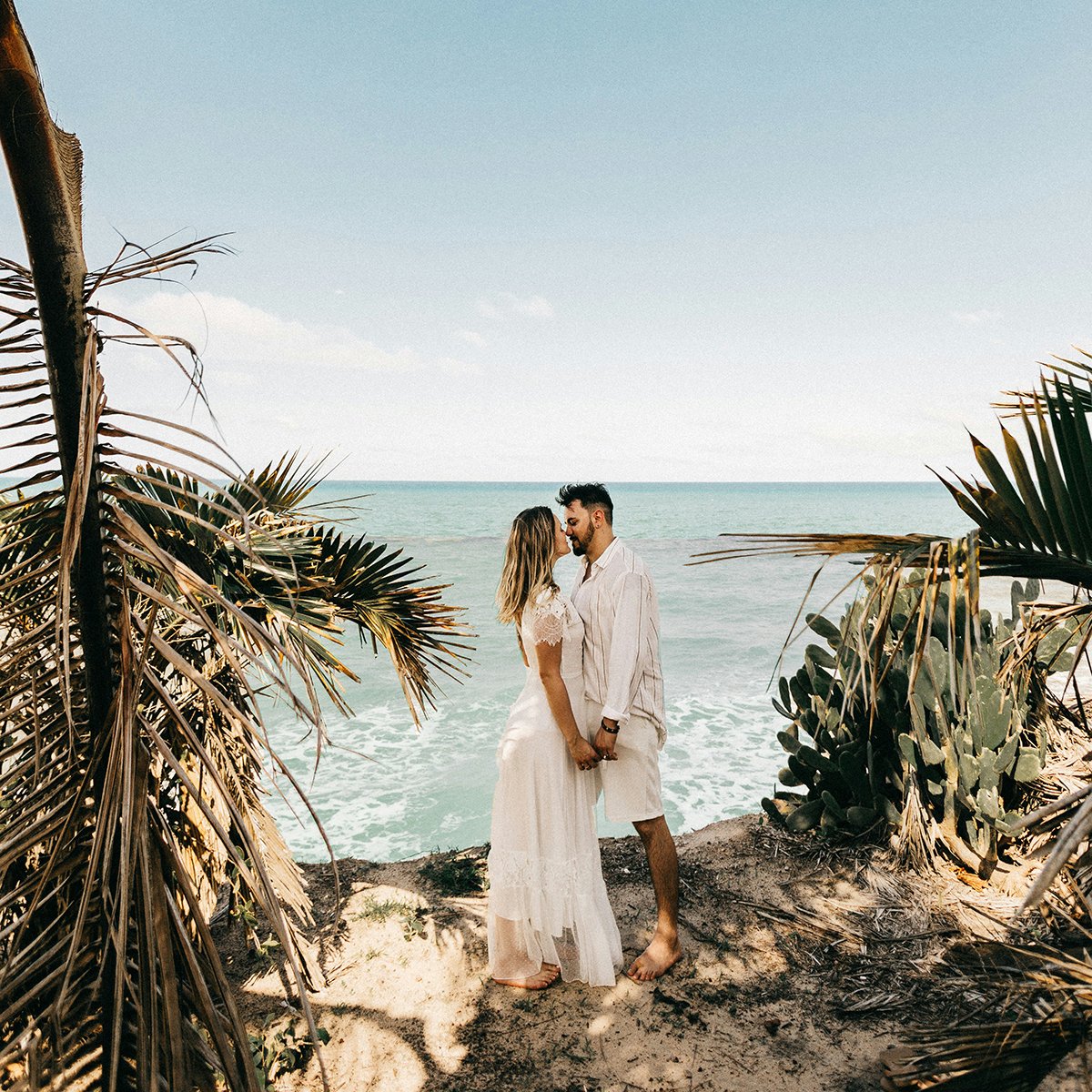 A couple, holding hands, stands on a beach with palm trees and the ocean surrounding them, creating a picturesque setting for a destination wedding.