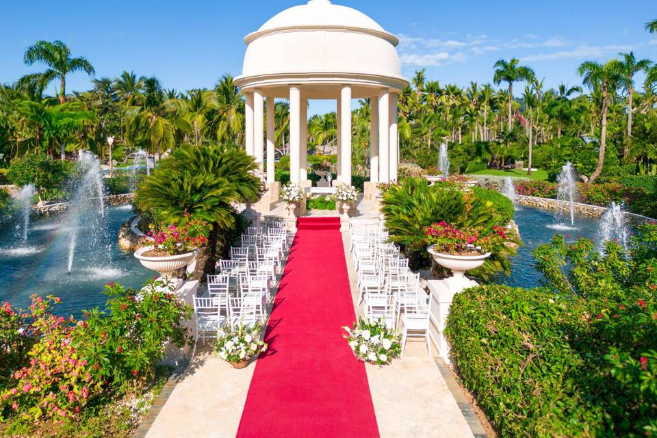 The outdoor wedding ceremony at Wyndham Alltra Punta Cana features a red carpet aisle surrounded by white chairs, fountains, and lush greenery, all leading to an elegant white dome structure.