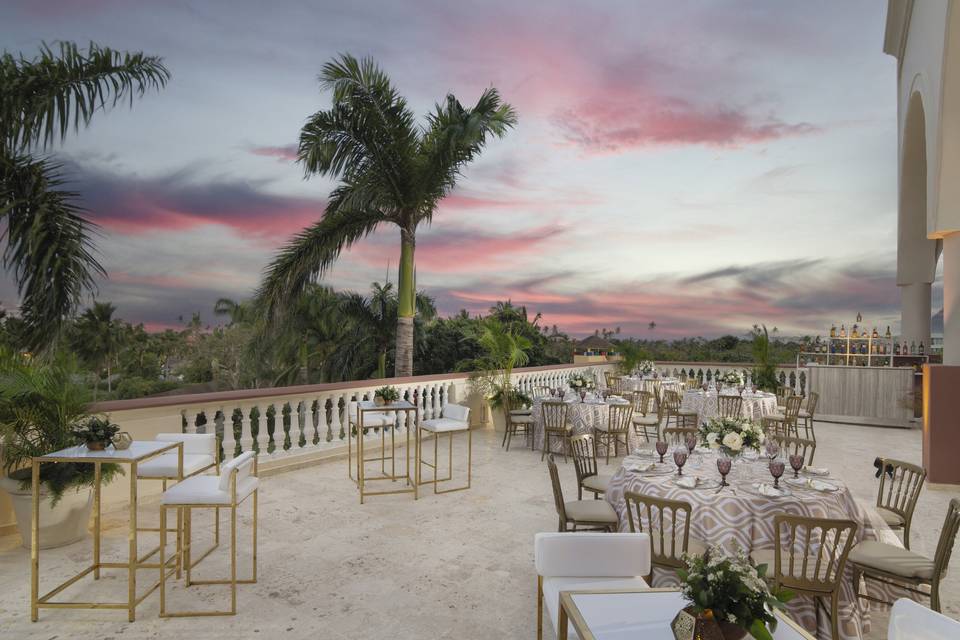A charming event setup awaits you at Wyndham Alltra Punta Cana, featuring elegantly arranged tables and chairs on the outdoor terrace, surrounded by palm trees and set against a vibrant sunset sky.