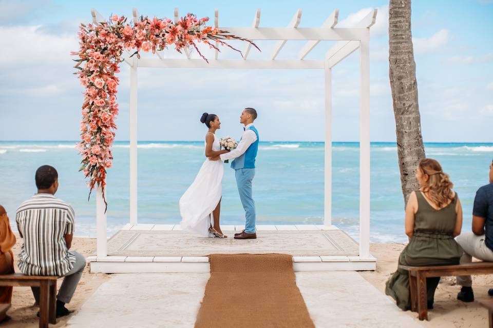 At Wyndham Alltra Punta Cana, a couple stands under a flower-adorned arch on the beach, exchanging vows as guests seated on benches face the ocean.