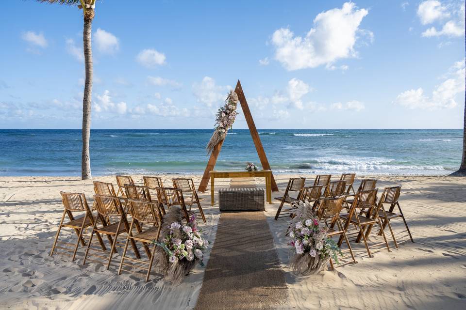 Enjoy a beach wedding setup at Wyndham Alltra Punta Cana featuring wooden chairs, a triangular flower-adorned altar, and a burlap aisle runner, all facing the ocean.