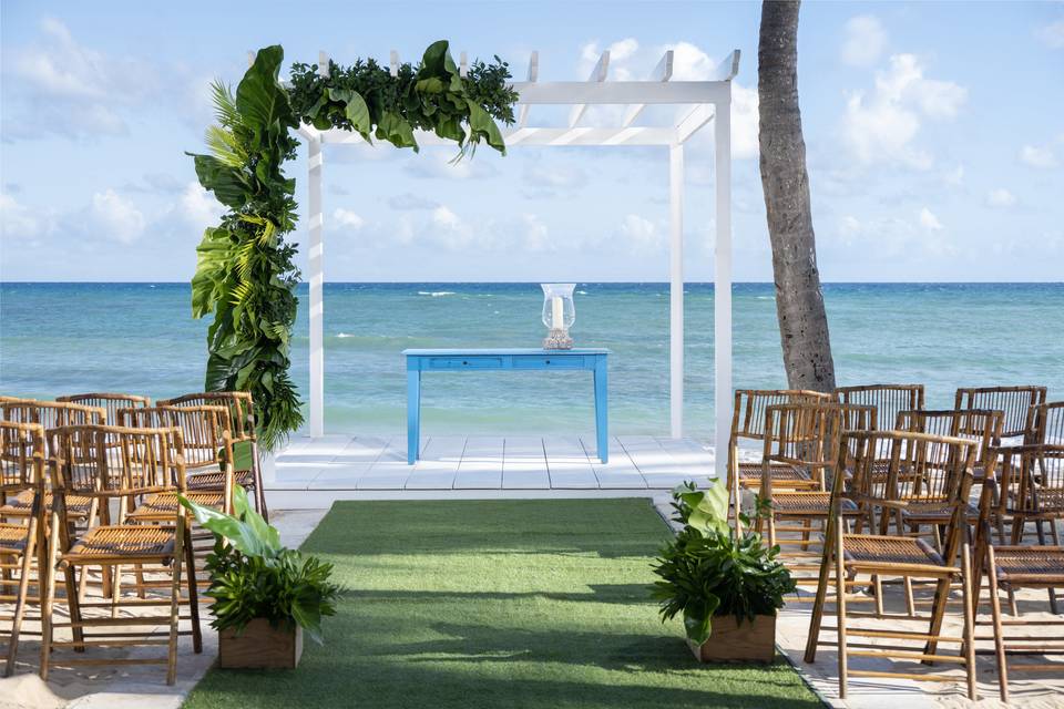 An outdoor beach wedding setup at Wyndham Alltra Punta Cana features a white pergola adorned with green foliage, and bamboo chairs facing the ocean. A blue table is elegantly placed under the pergola.
