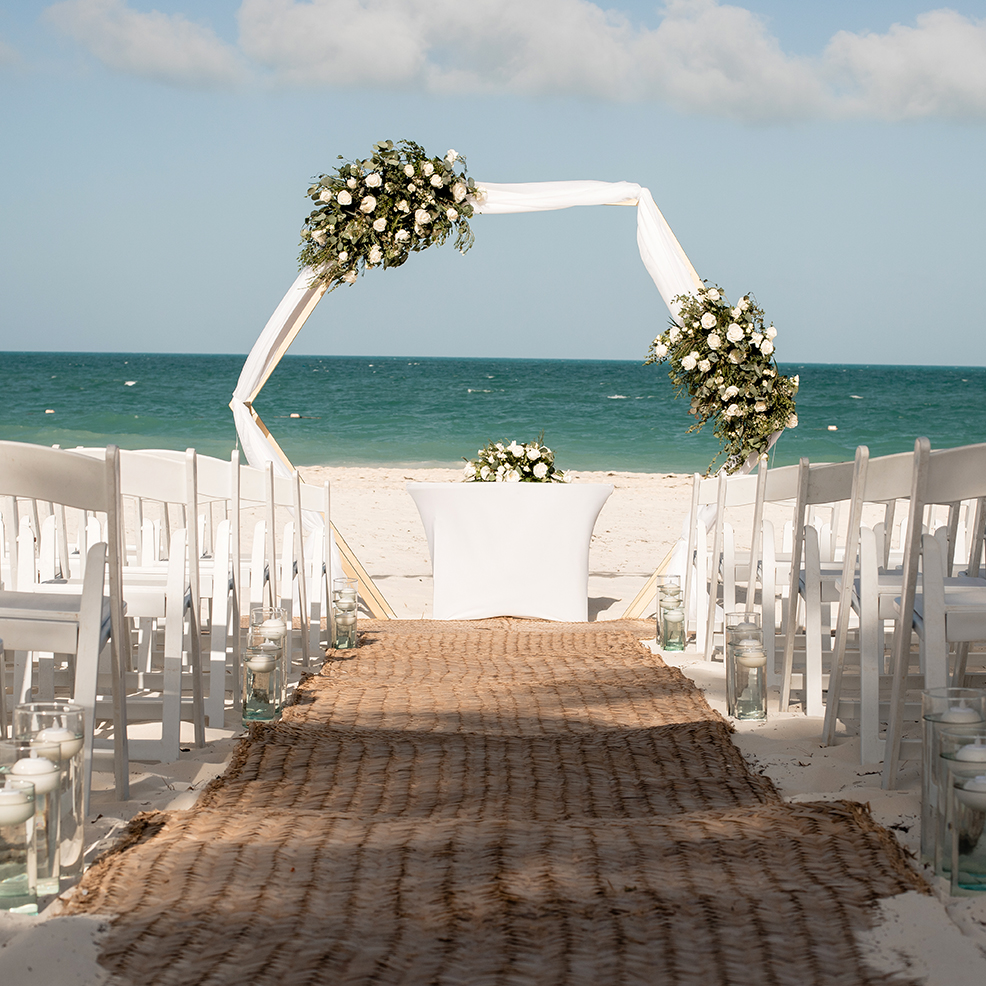 A destination wedding beautifully arranged with a floral arch, white chairs, and an aisle gracefully leading to the ocean.