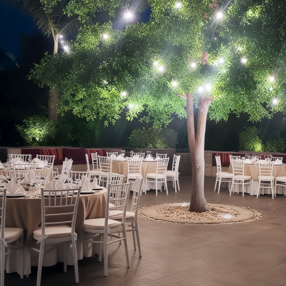 A picturesque destination wedding setup featuring round tables adorned with elegant white chairs under a beautifully lit tree, twinkling with enchanting string lights.