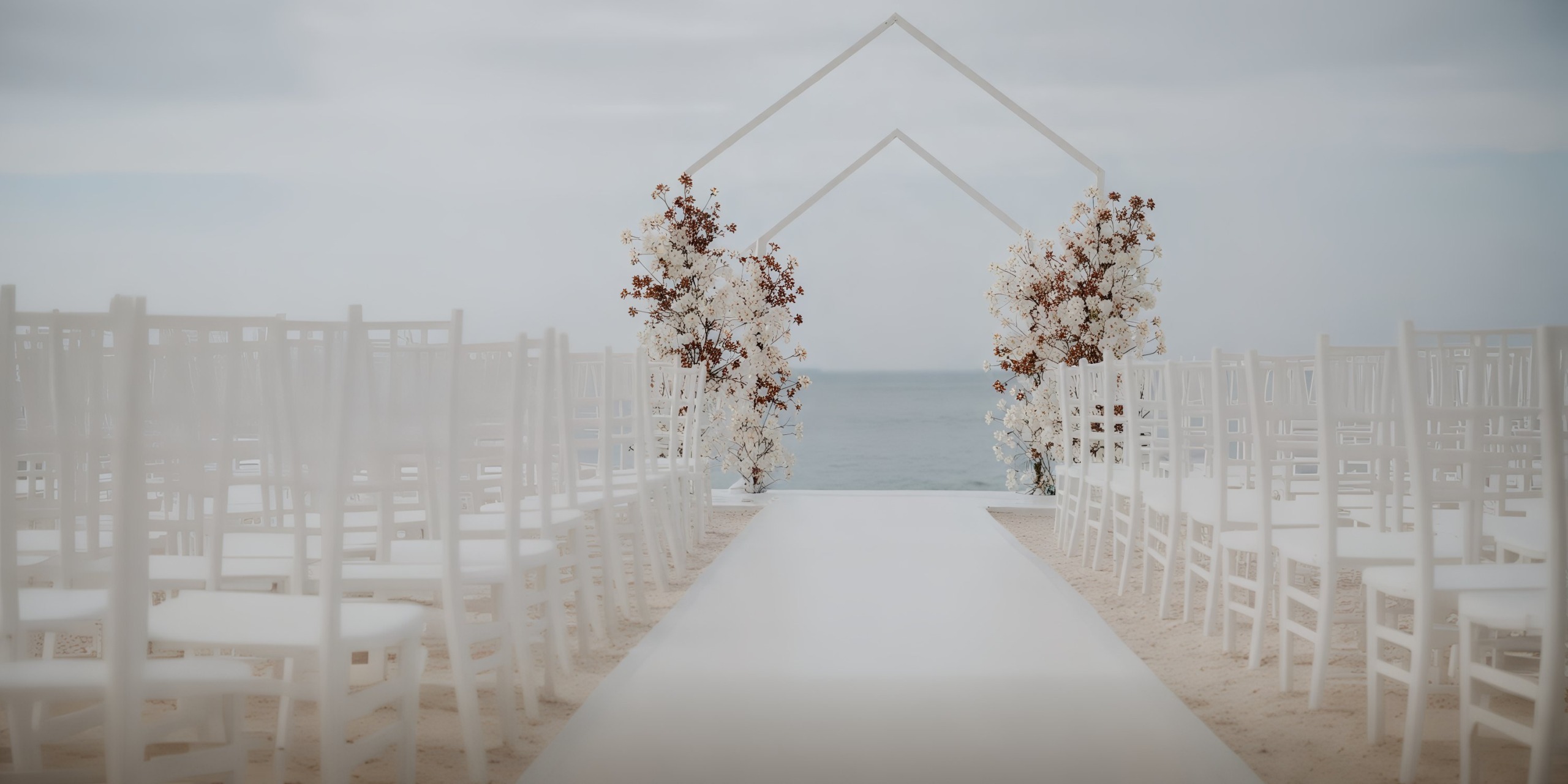A picturesque destination wedding aisle by the sea, featuring elegant white chairs and a beautifully adorned floral arch.