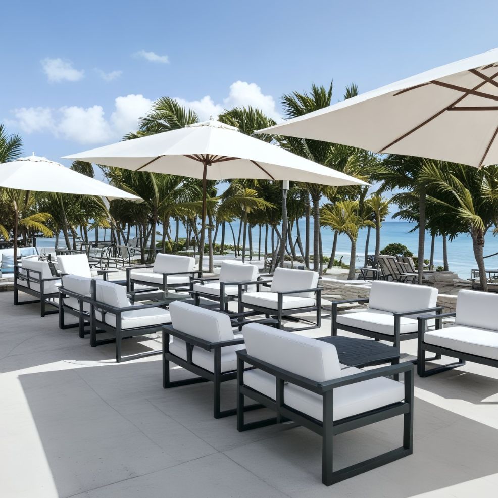 A picturesque outdoor patio, perfect for a destination wedding, features white chairs, tables, and umbrellas nestled among palm trees with a breathtaking ocean backdrop.