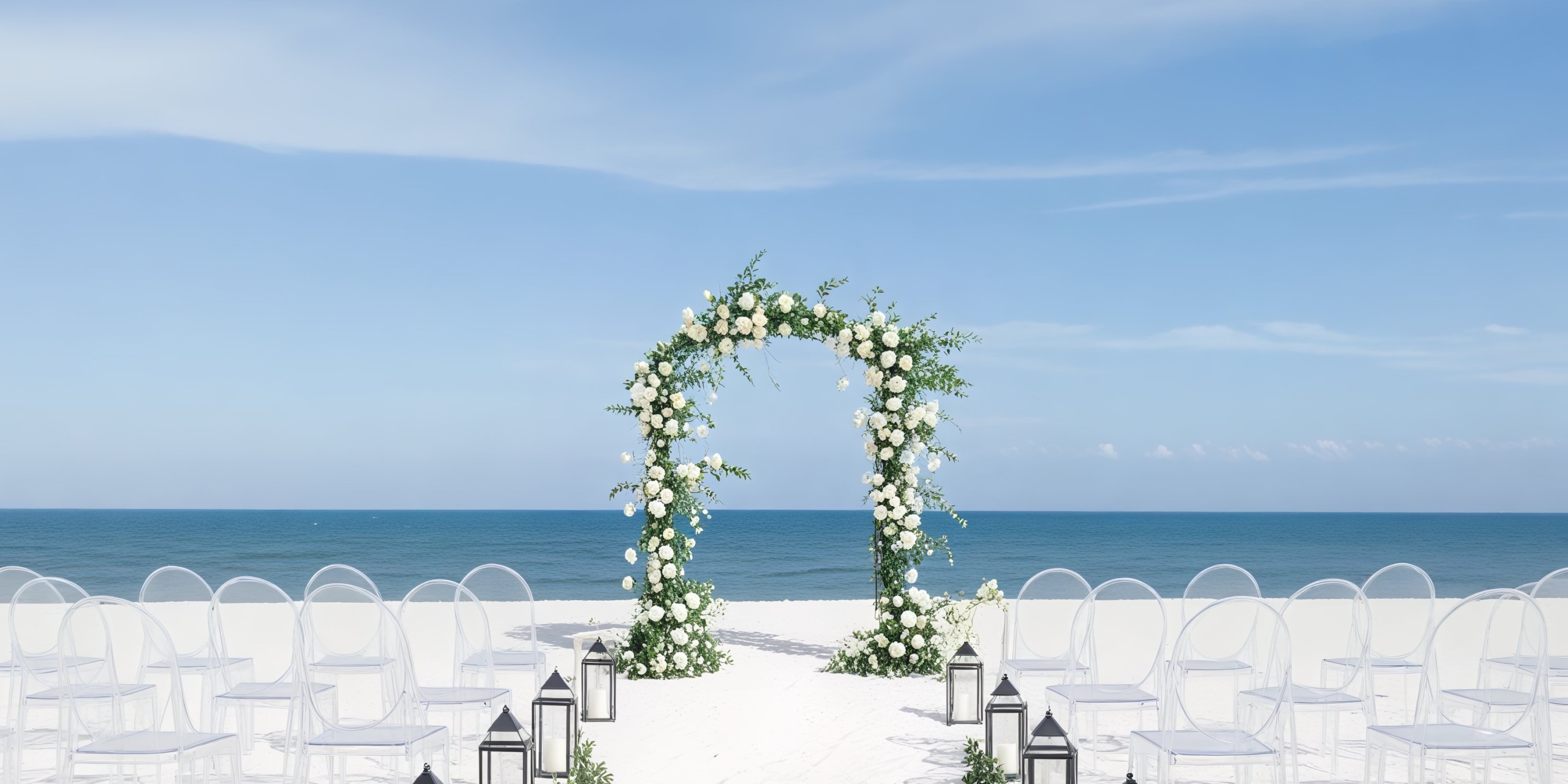 A beautiful destination wedding setup featuring a floral arch, chairs, and lanterns all oriented towards the ocean.