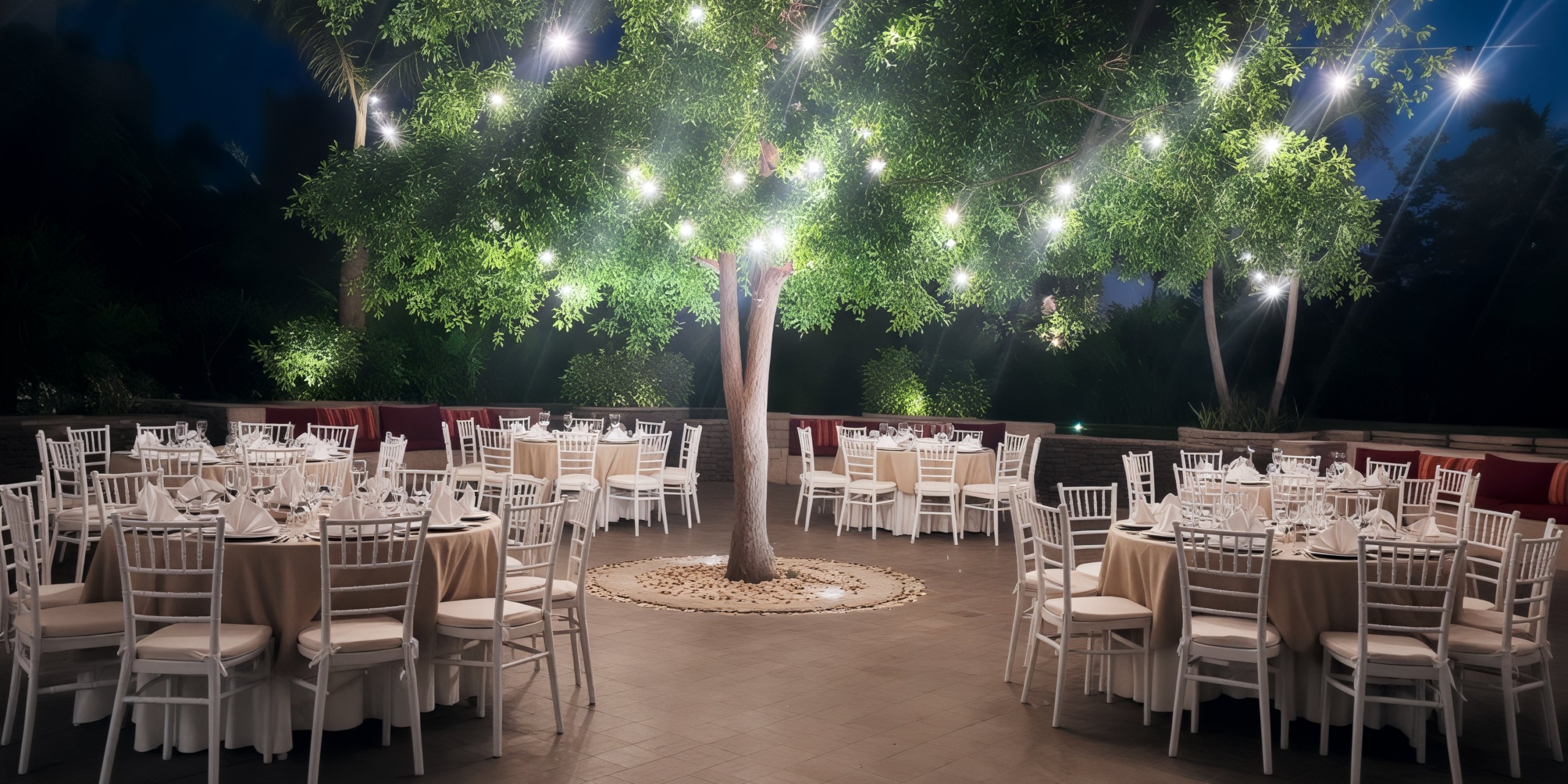 Wedding-themed outdoor dining area featuring round tables, elegant white chairs, and a beautiful tree decorated with string lights.