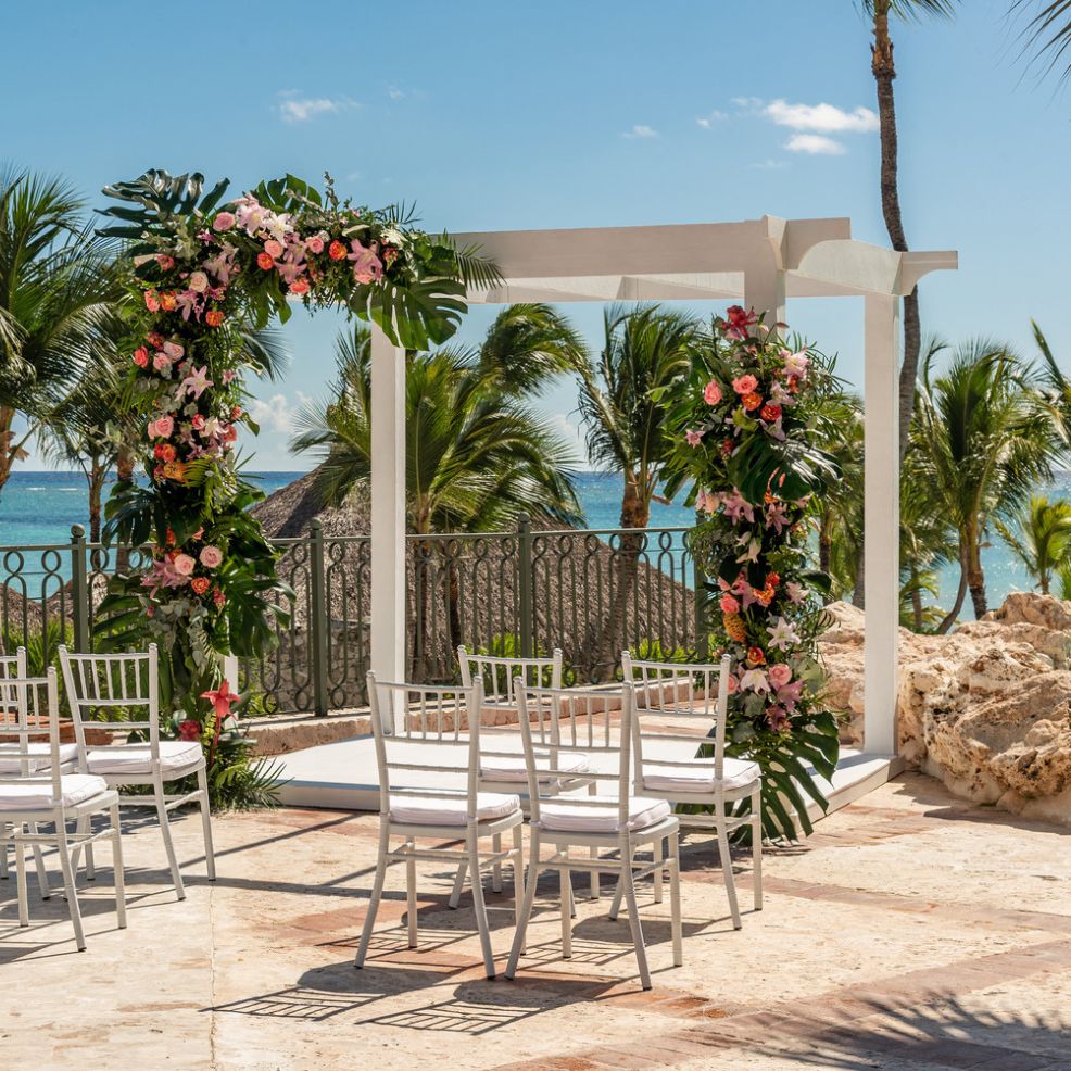 A destination wedding setup featuring an outdoor arch adorned with flowers, surrounded by white chairs, palm trees, and offering a stunning ocean view.
