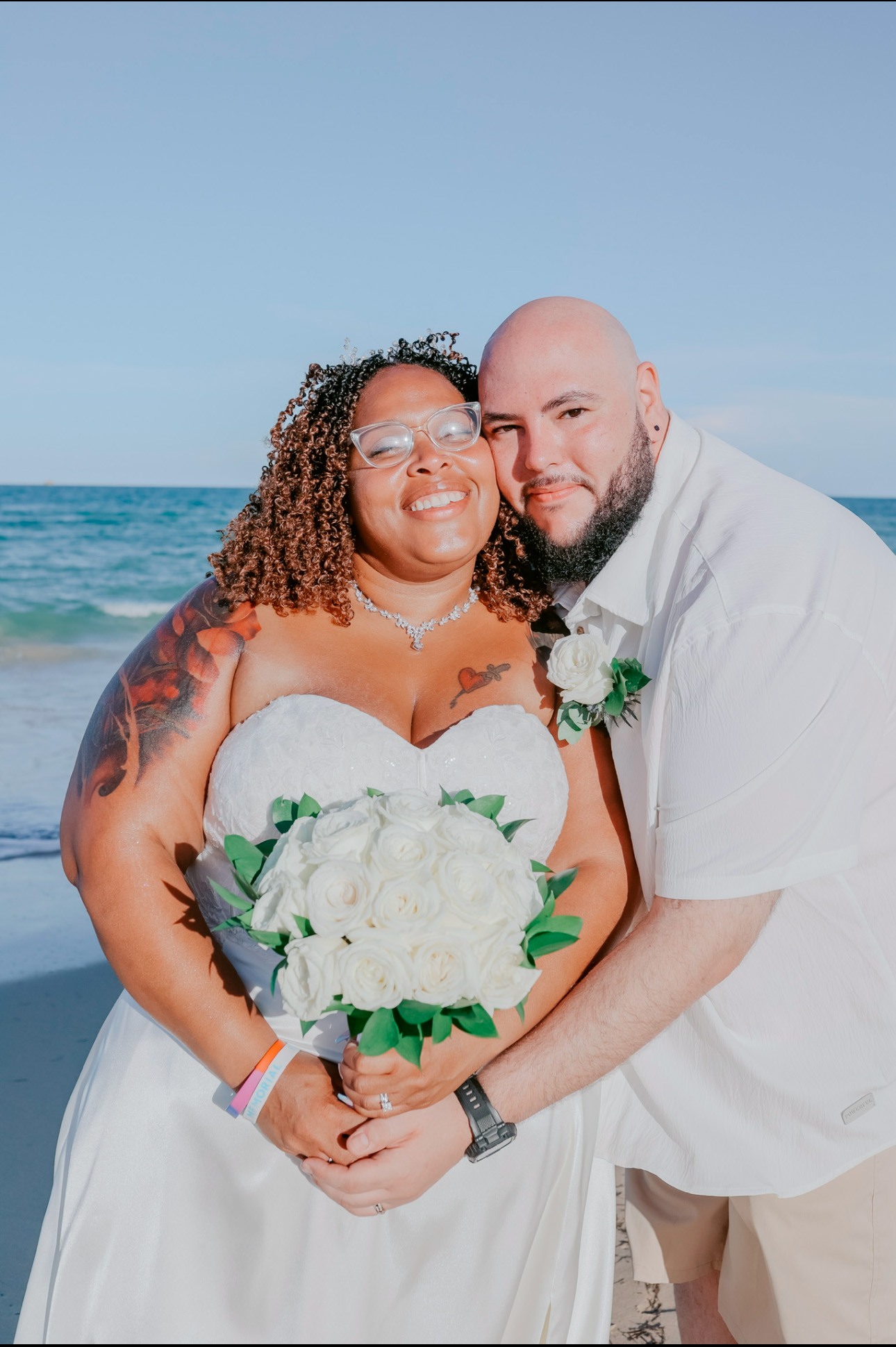 A couple in wedding attire beams with joy on a beach, holding a bouquet of white roses, perfectly capturing the essence of a destination wedding.