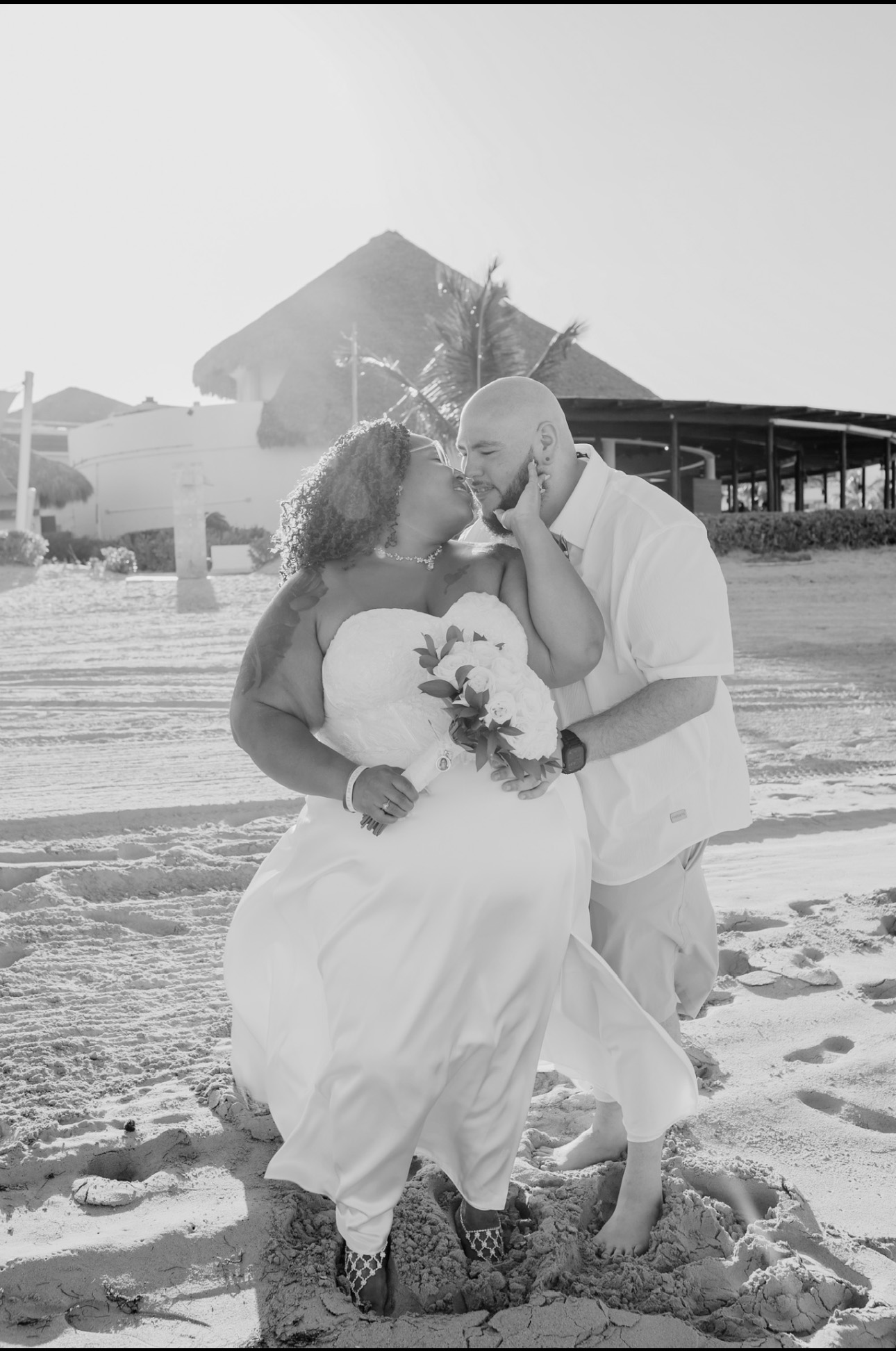 A couple in wedding attire embraces and kisses on a sandy beach, capturing a beautiful moment at their destination wedding.