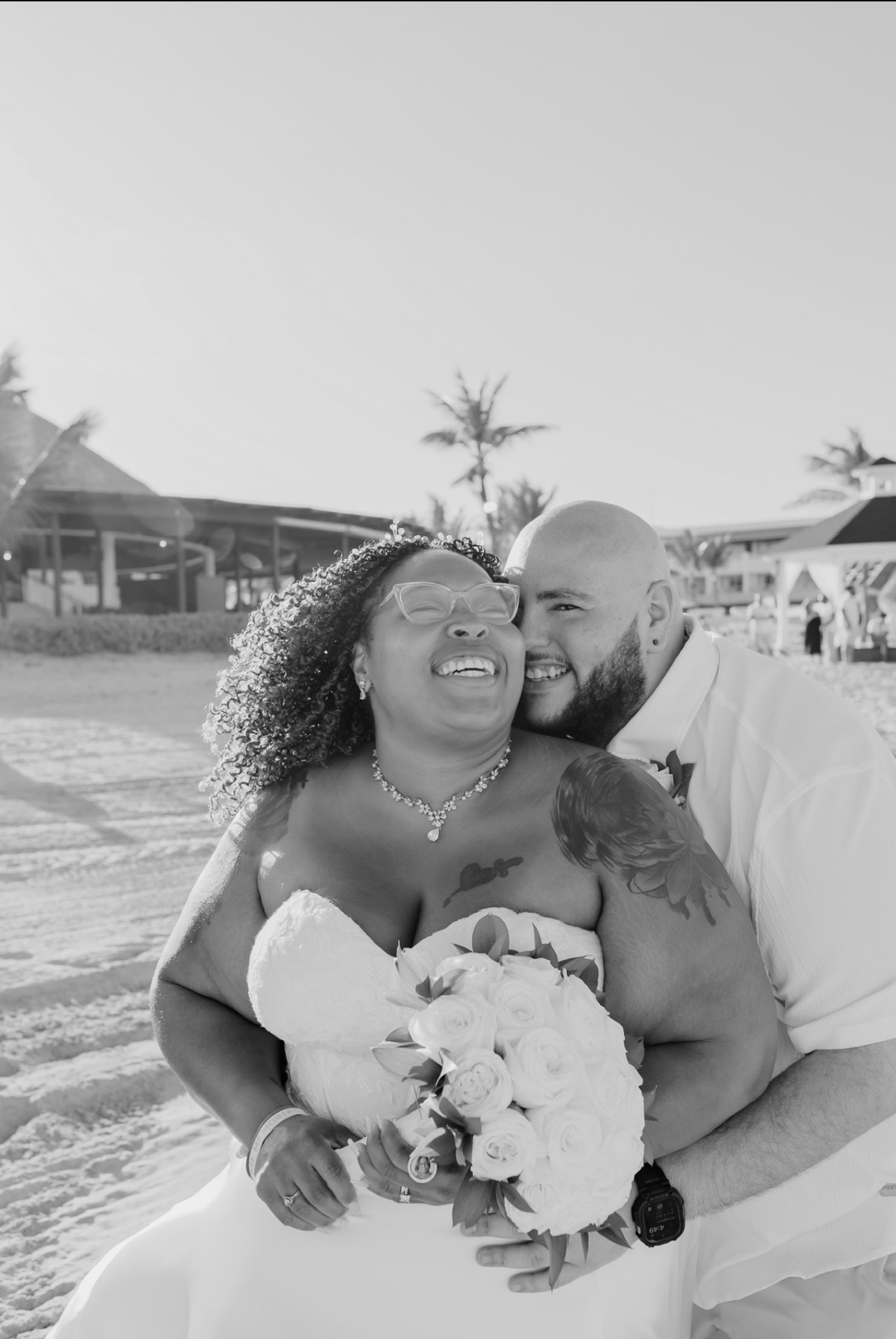 The bride and groom are beaming with joy on a beach, holding a bouquet of roses, creating the perfect scene for their destination wedding.