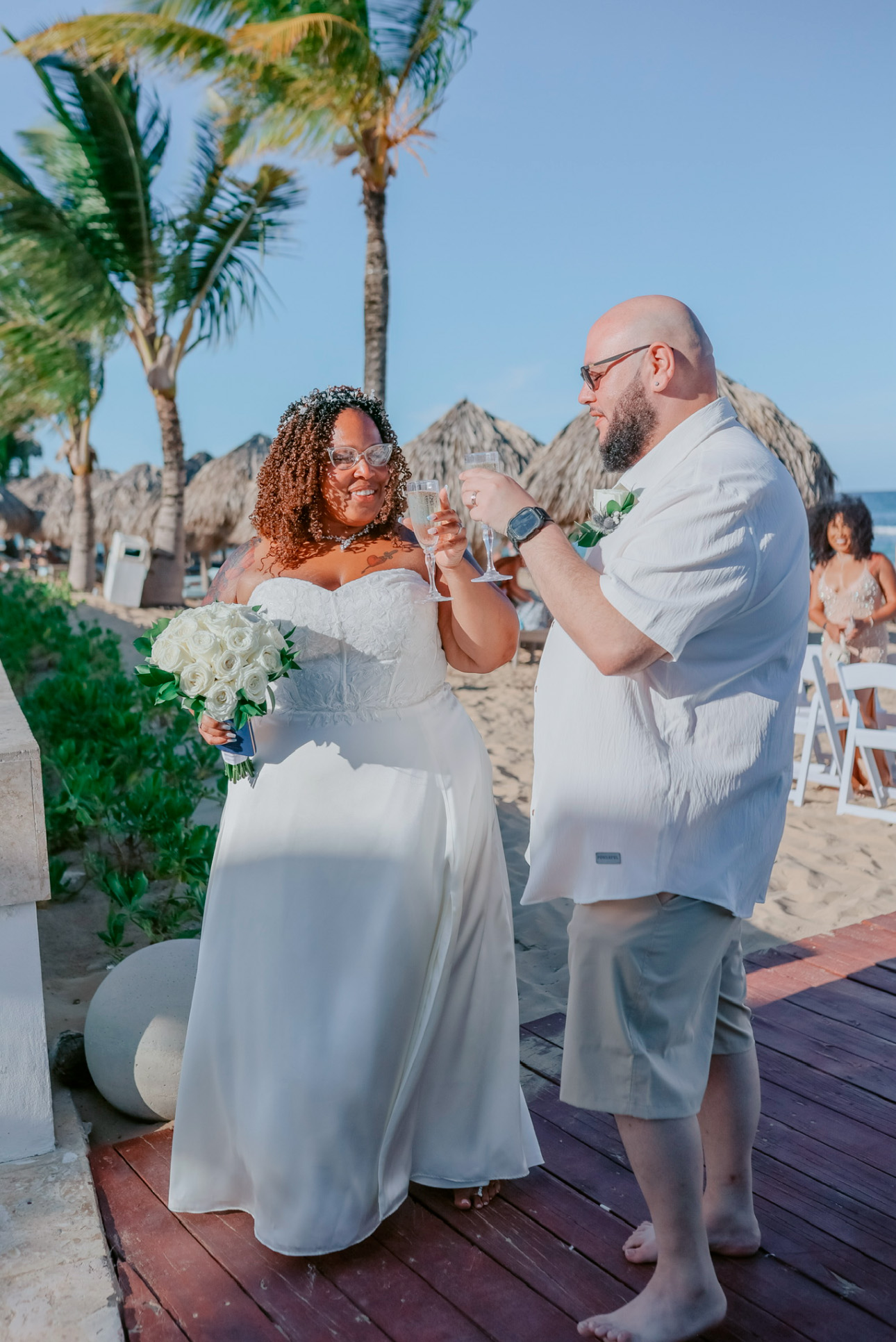 The bride and groom clink their glasses on a tropical beach, celebrating their destination wedding.