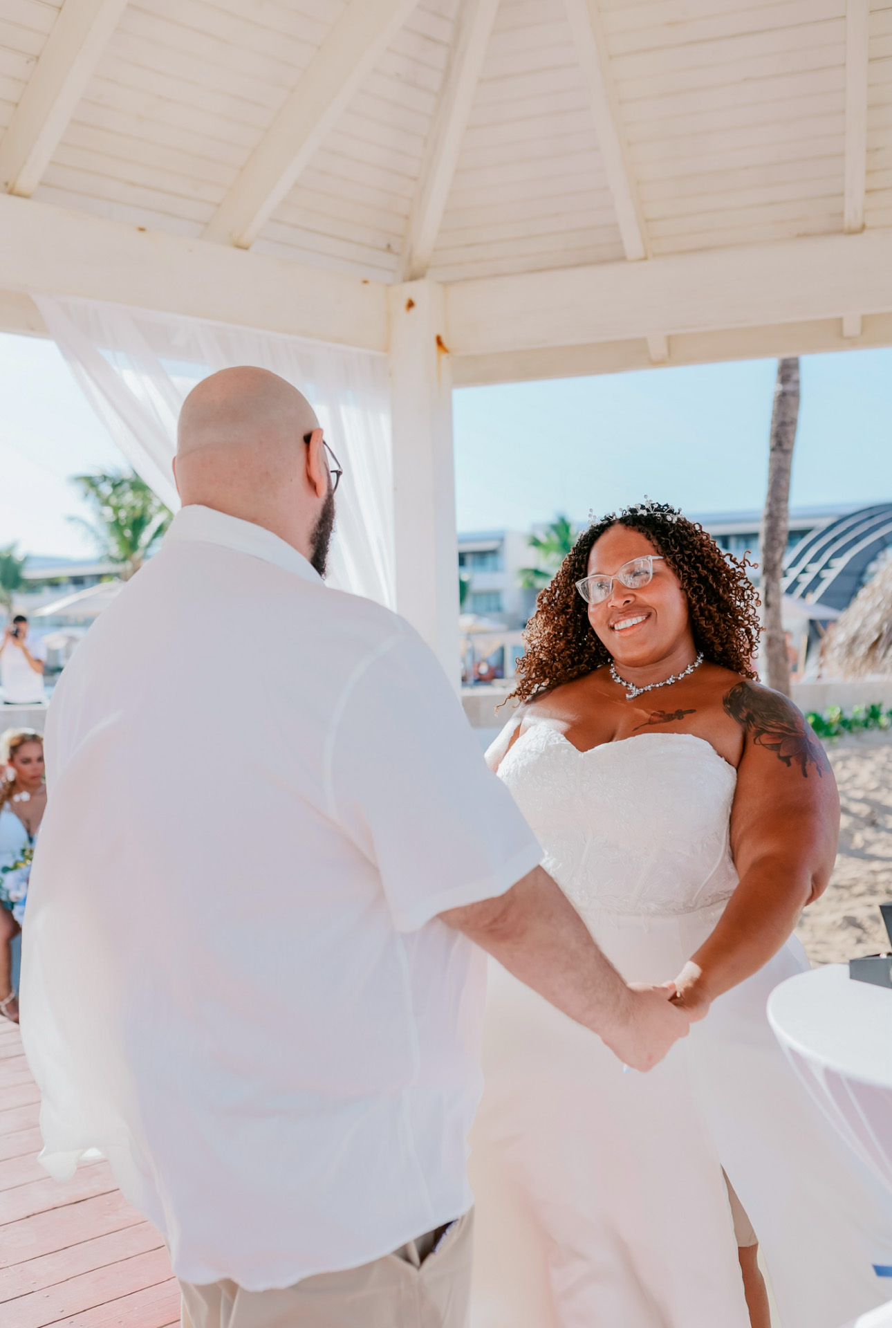 A couple dressed in elegant white attire holds hands during an outdoor destination wedding ceremony.