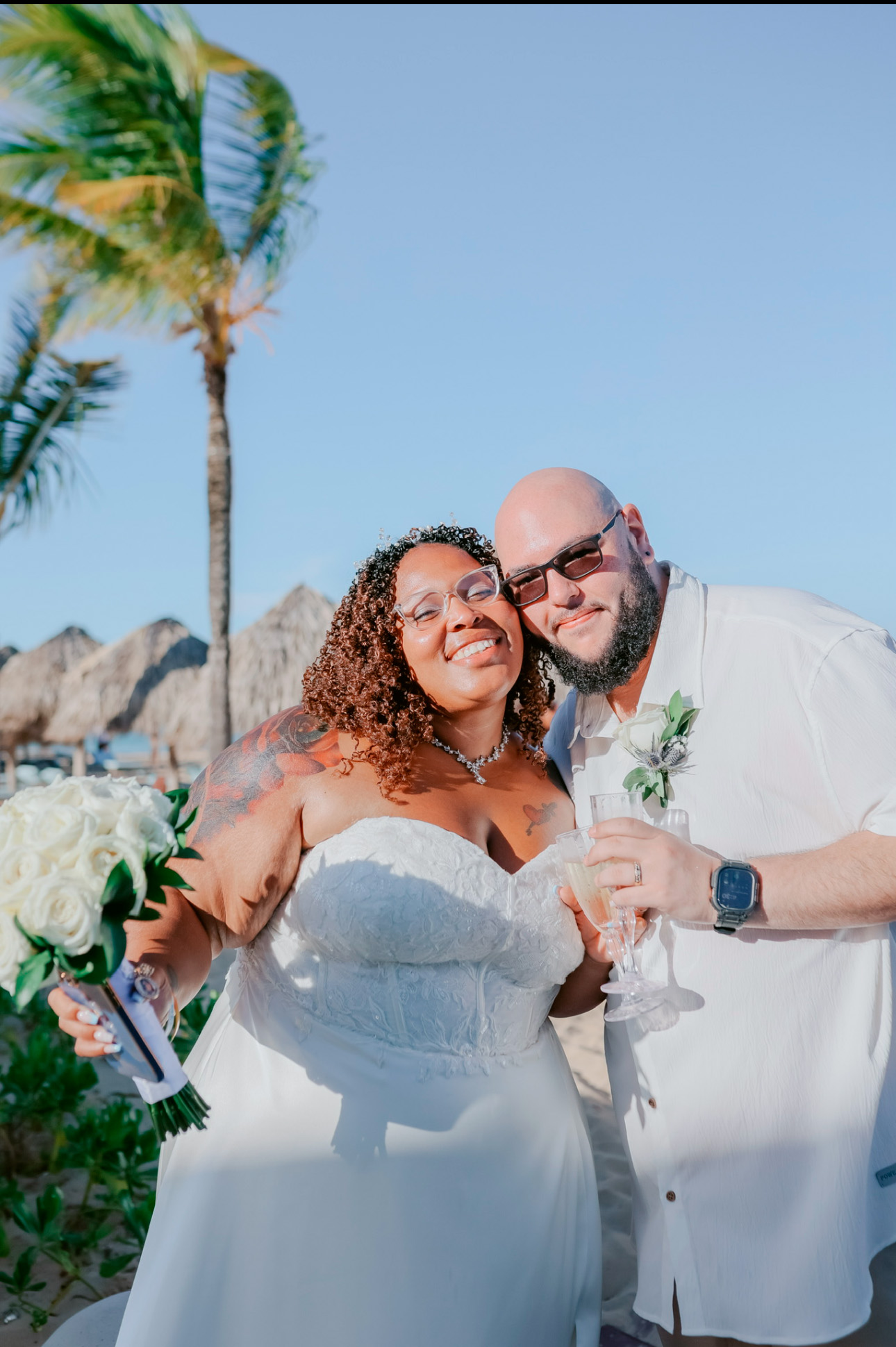 A couple dressed in wedding attire poses with drinks on a sunny beach, creating the perfect destination wedding moment.