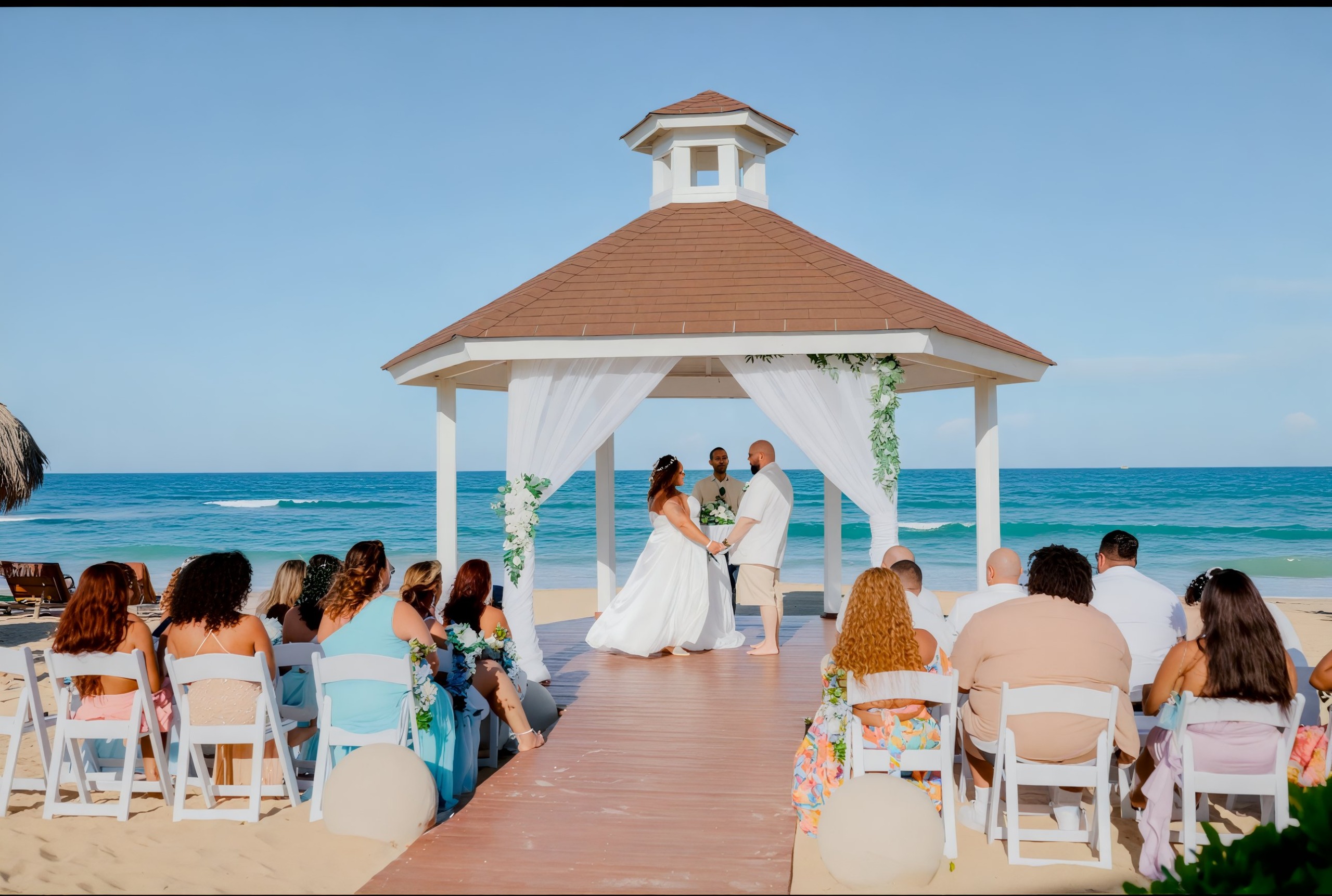 A destination wedding ceremony takes place under a charming gazebo on the beach, where guests are seated in rows, offering a stunning view of the ocean.