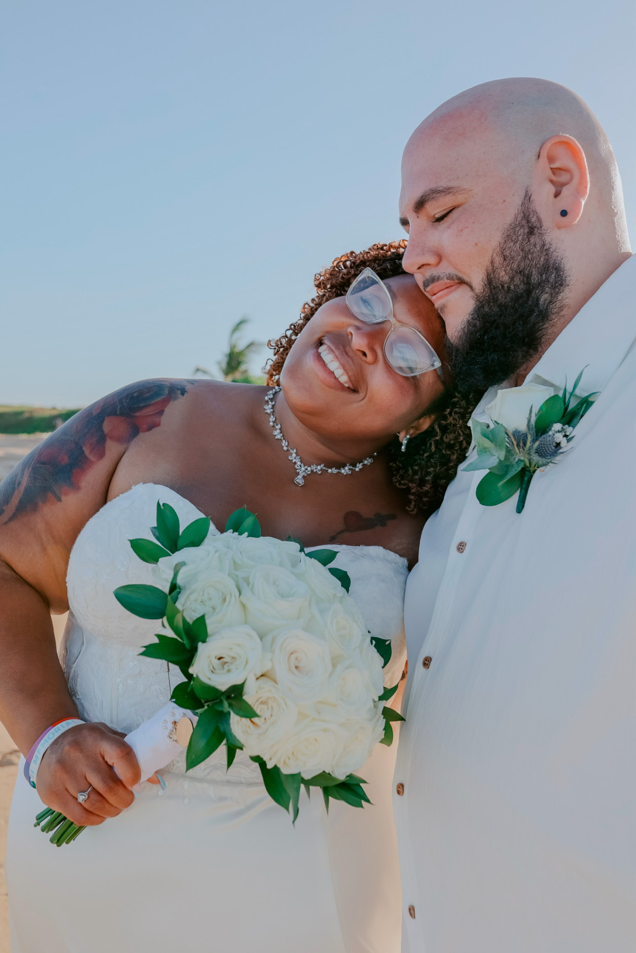 The bride and groom beam with joy as they embrace on a sunny day, holding a bouquet of white roses at their destination wedding.