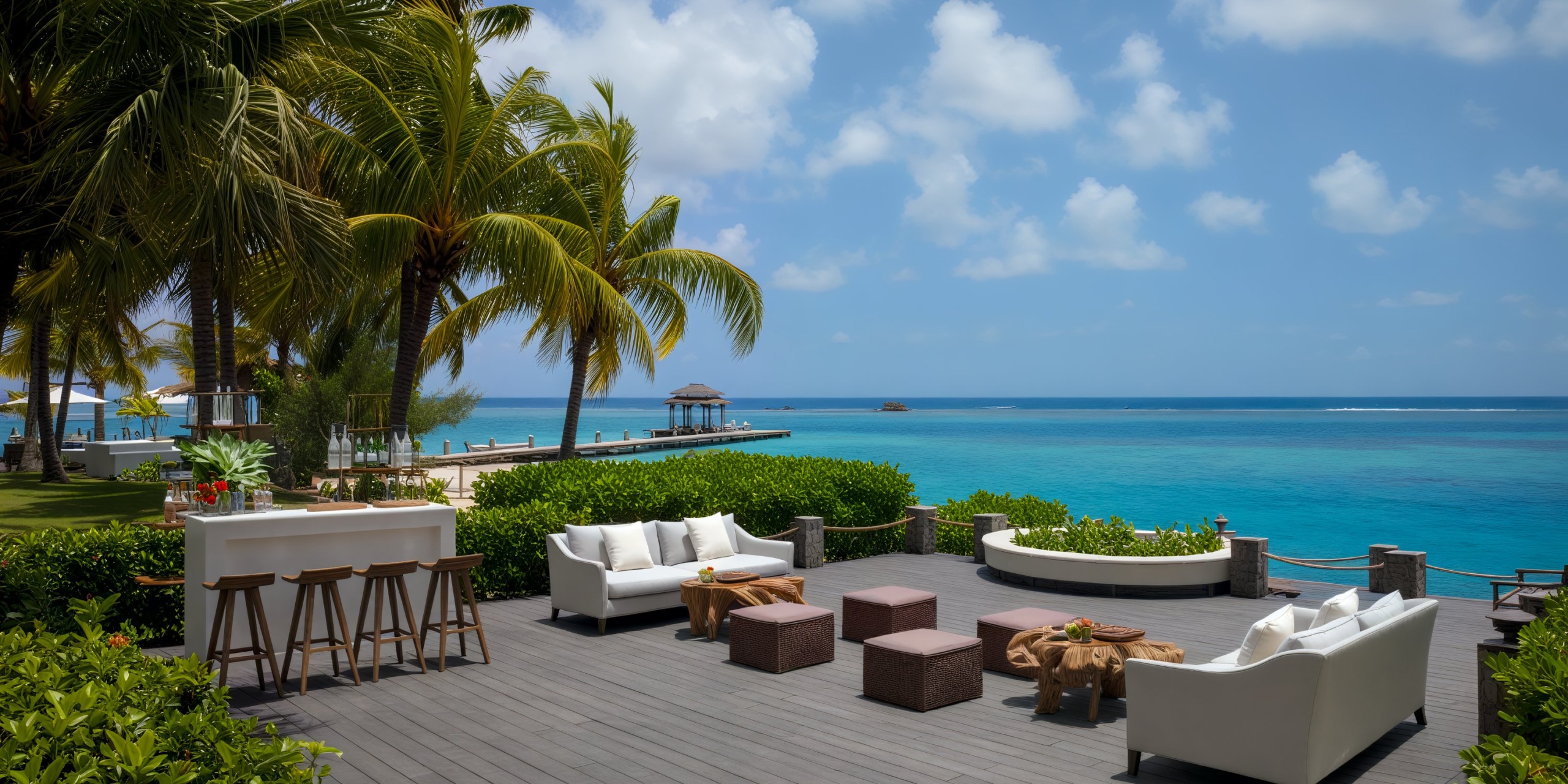 A beachfront patio, adorned with white sofas and bar stools, surrounded by palm trees and offering stunning ocean views, makes for a perfect destination wedding venue.