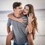 A woman joyfully displays her ring while piggybacking on a man during their destination wedding at the beach.