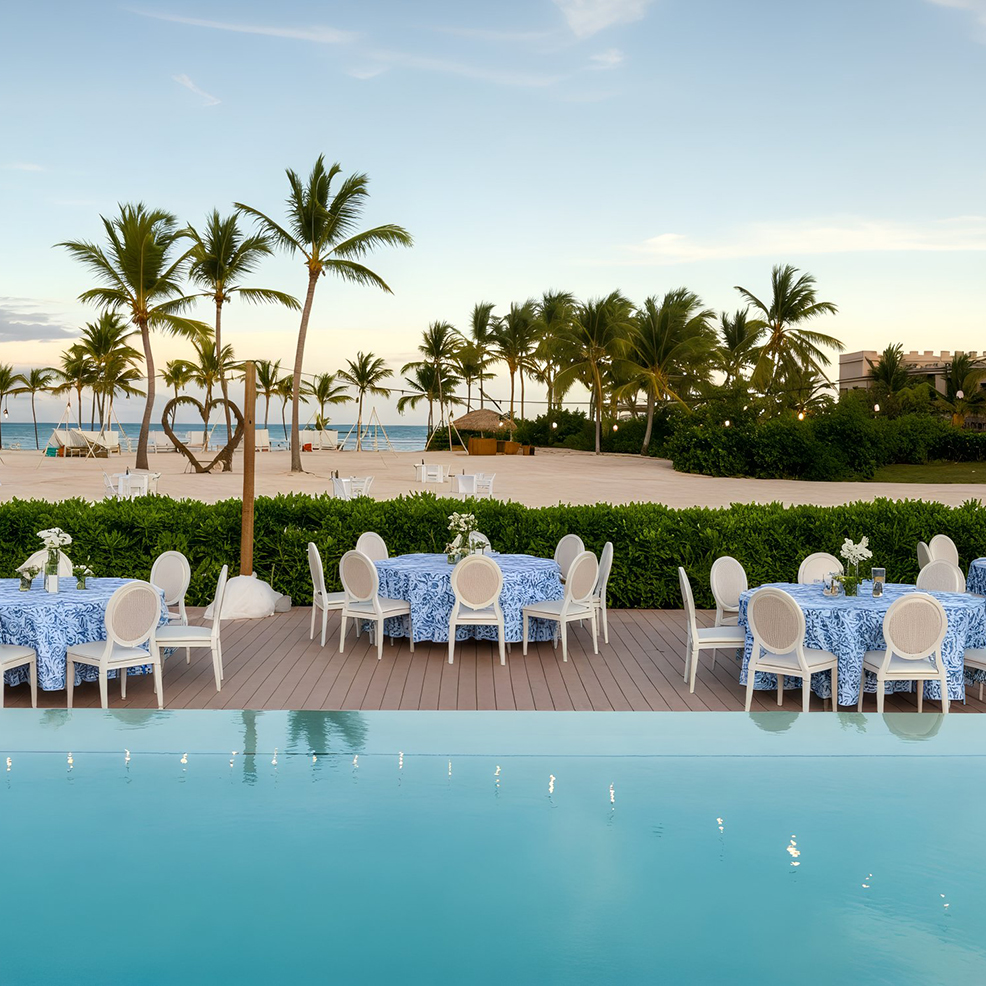 An outdoor dining arrangement set by a pool, enhanced with palm trees and a beach backdrop, ideal for a destination wedding.