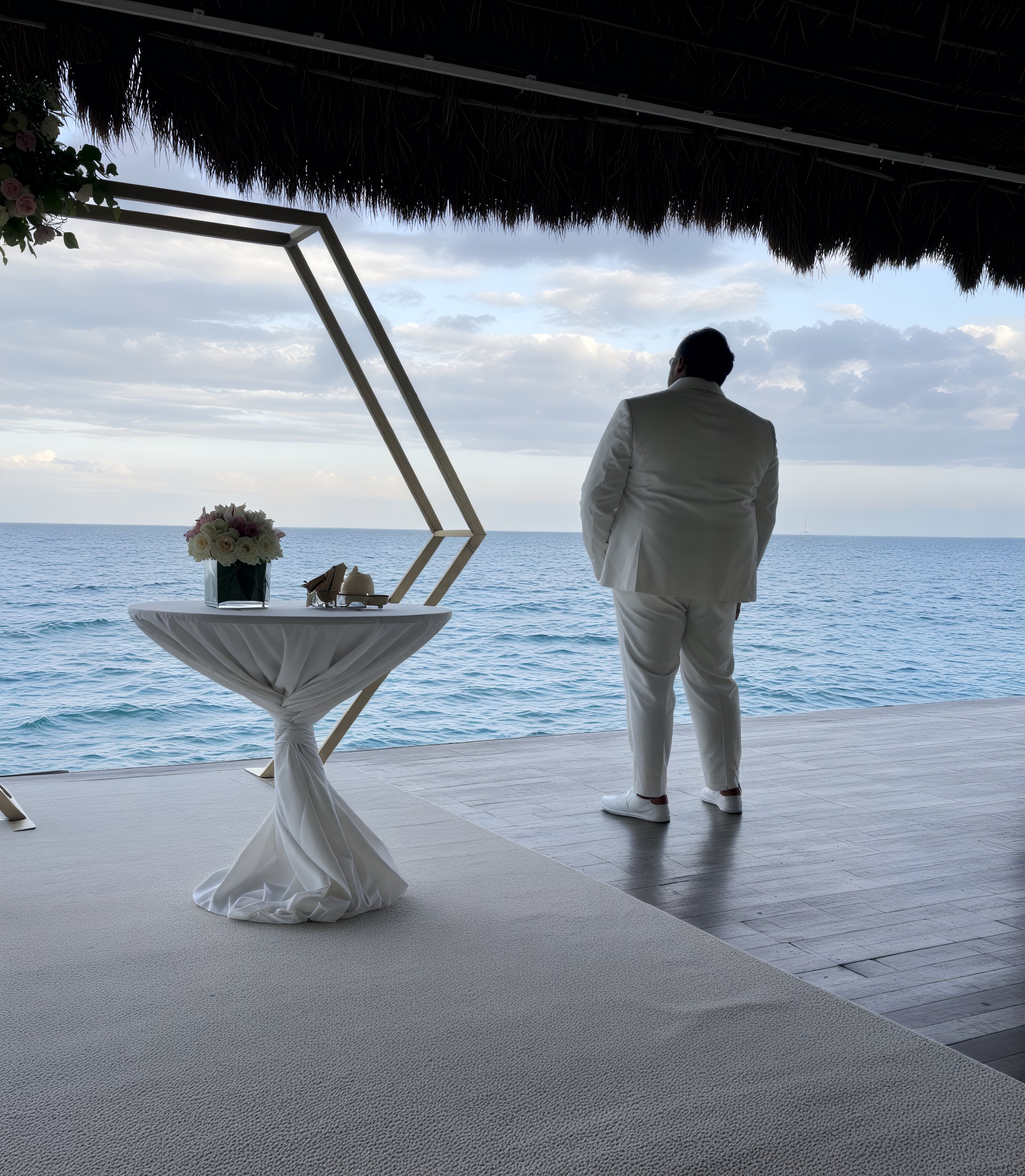 A person in a white suit stands alone on a deck at the destination wedding, with Sheneka and Andre gazing at the ocean.