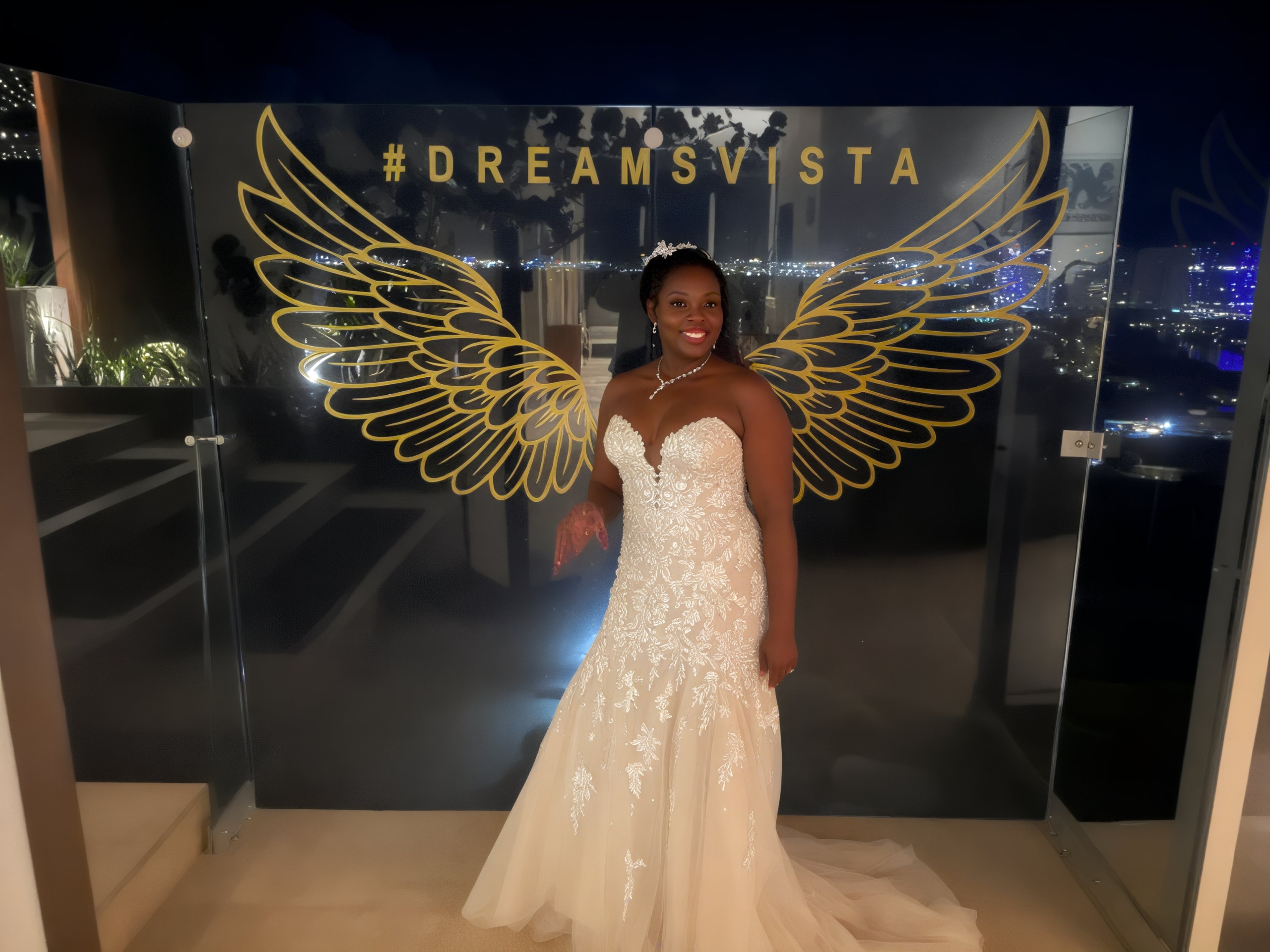The bride, adorned in Sheneka and Andre's elegant gown, poses gracefully beside a glass panel, accentuated by golden wings and the shimmering city lights—a true vision of a destination wedding.