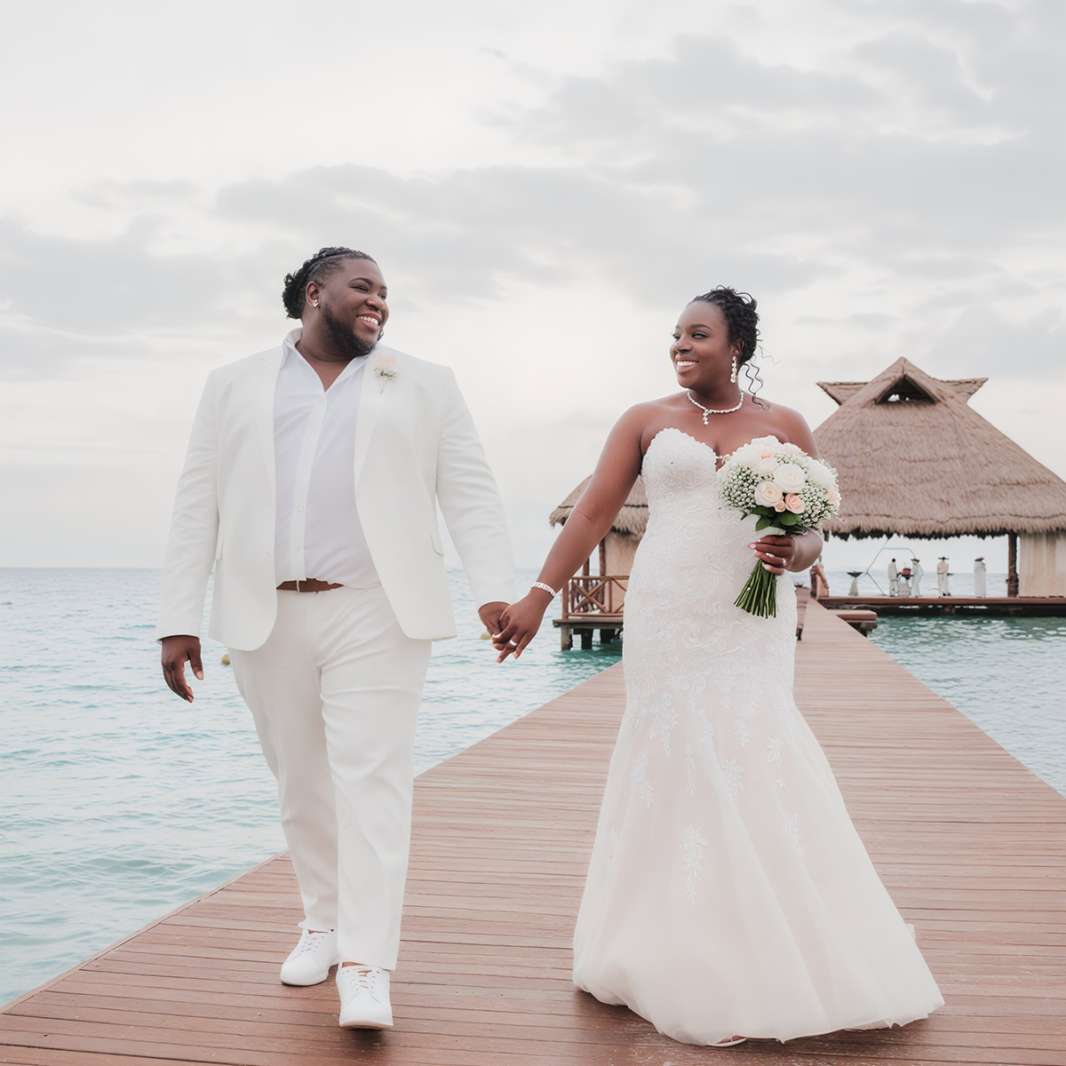 Sheneka and Andre, dressed in elegant white attire, hold hands on a seaside dock, creating a picturesque moment at their destination wedding by the ocean.