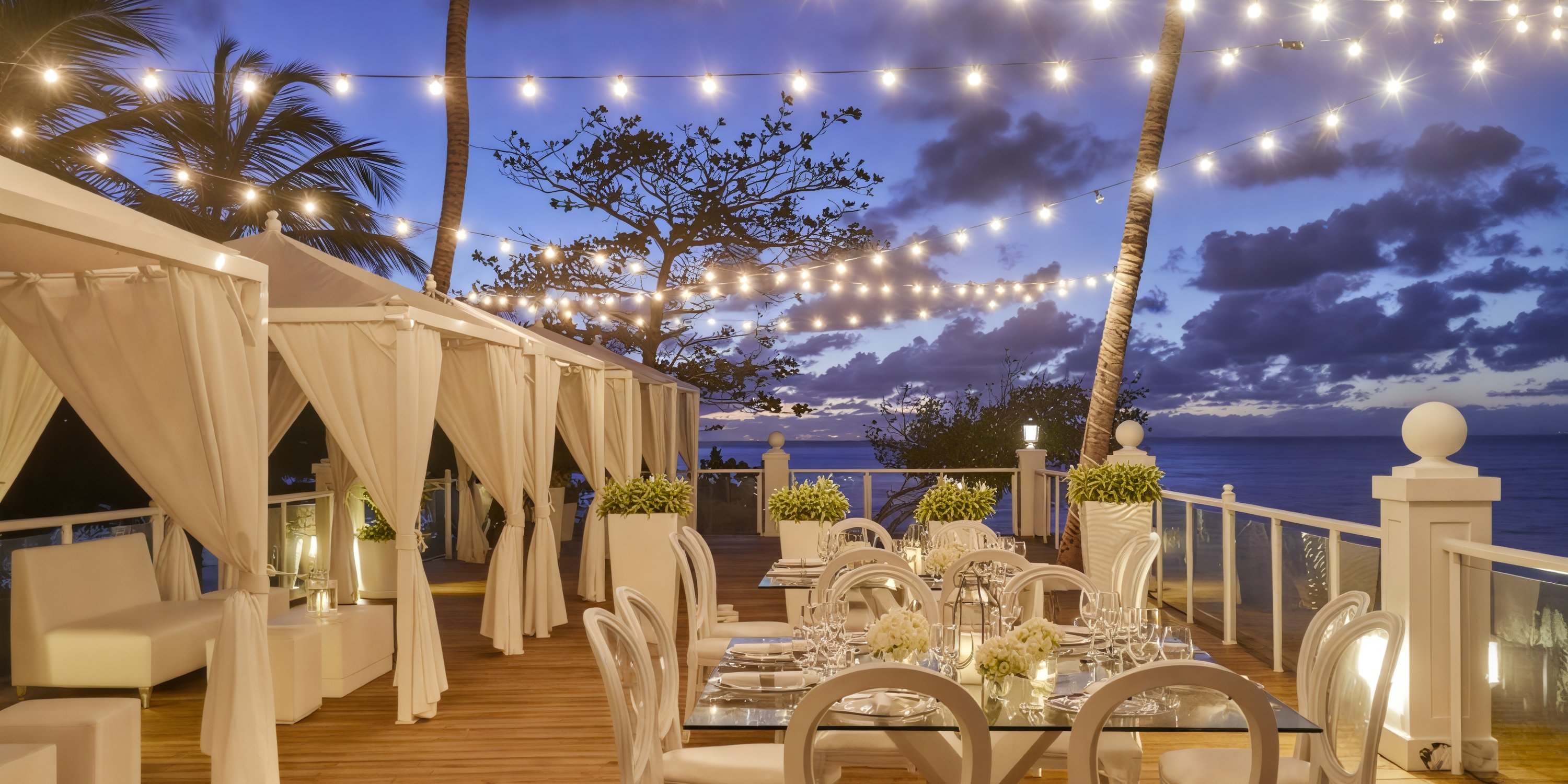 A sophisticated outdoor dining area, perfect for a destination wedding, adorned with string lights and offering breathtaking ocean views at dusk.
