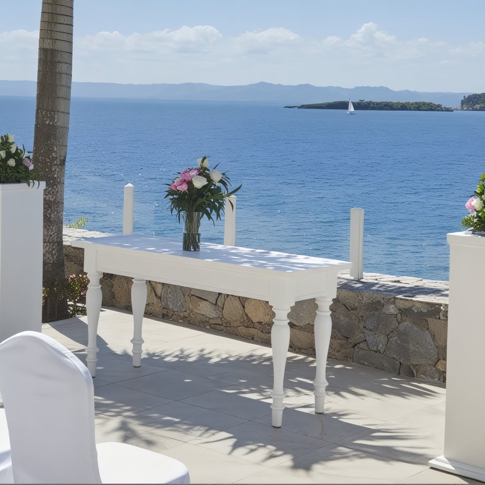 A white table adorned with a floral vase graces the seaside terrace, creating an idyllic setting for a destination wedding. In the distance, a sailboat glides gracefully under the expansive blue sky.