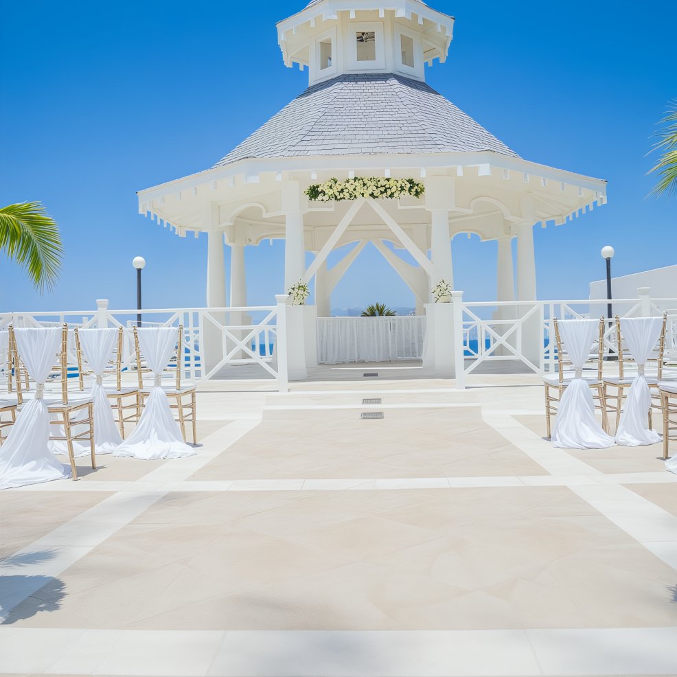 An outdoor wedding gazebo adorned with white chairs is set beneath a clear blue sky, perfect for a picturesque destination wedding.