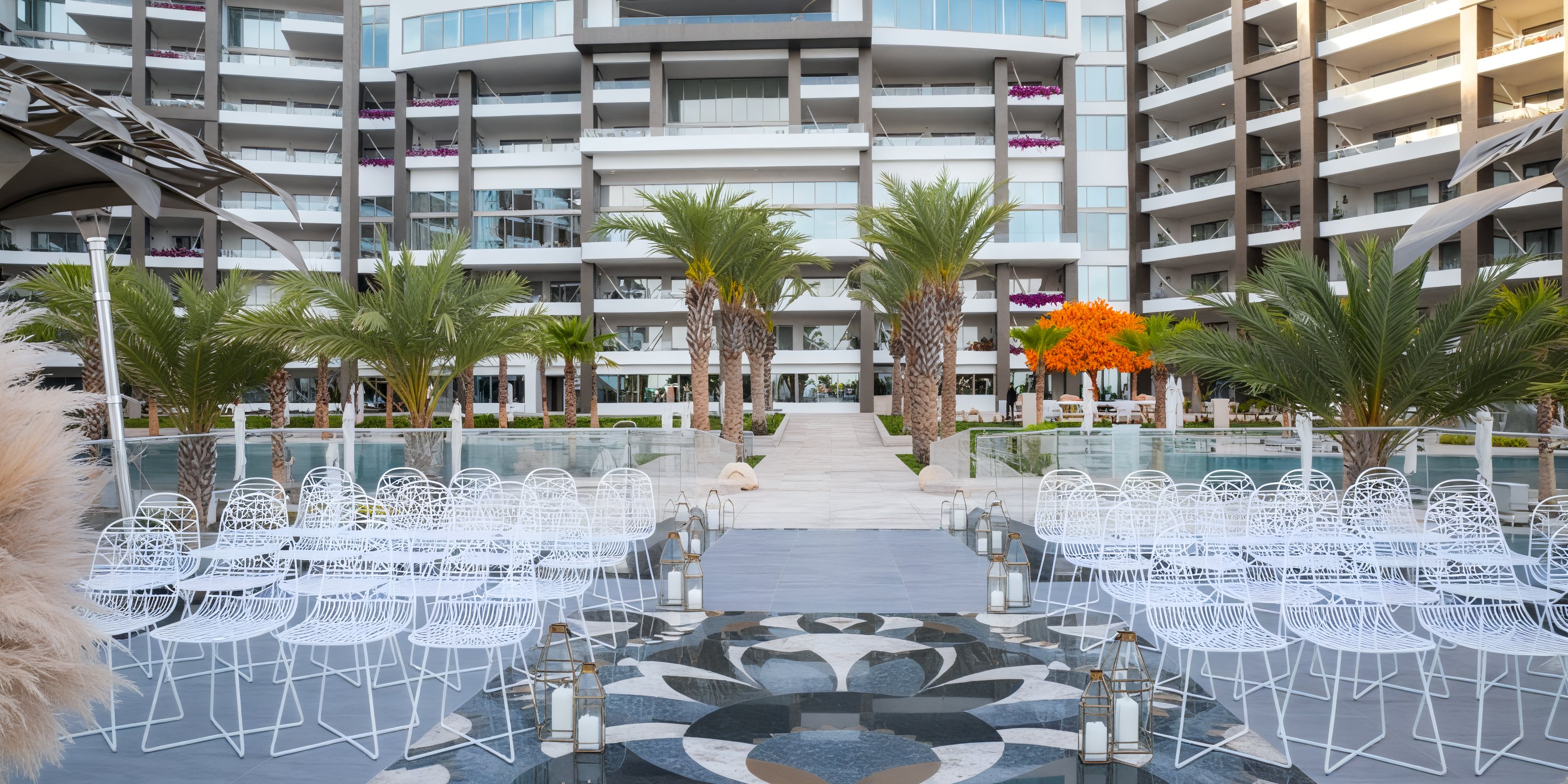 Experience a stunning destination wedding at Garza Blanca Los Cabos, where elegant white chairs are arranged amidst swaying palm trees and surrounded by the sleek lines of a modern building.