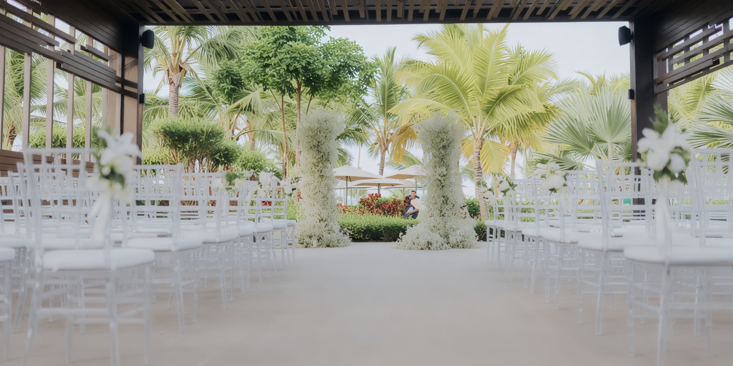 An idyllic setting for a destination wedding, featuring an outdoor aisle lined with white chairs and elegant floral arrangements, surrounded by lush tropical trees in the backdrop.