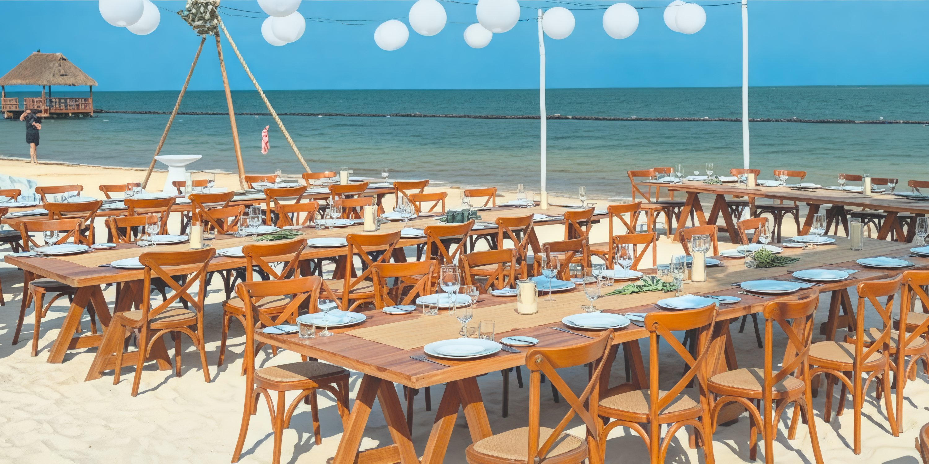 Tables adorned with beachfront elegance are set for a destination wedding, complemented by the enchanting glow of white hanging lanterns.