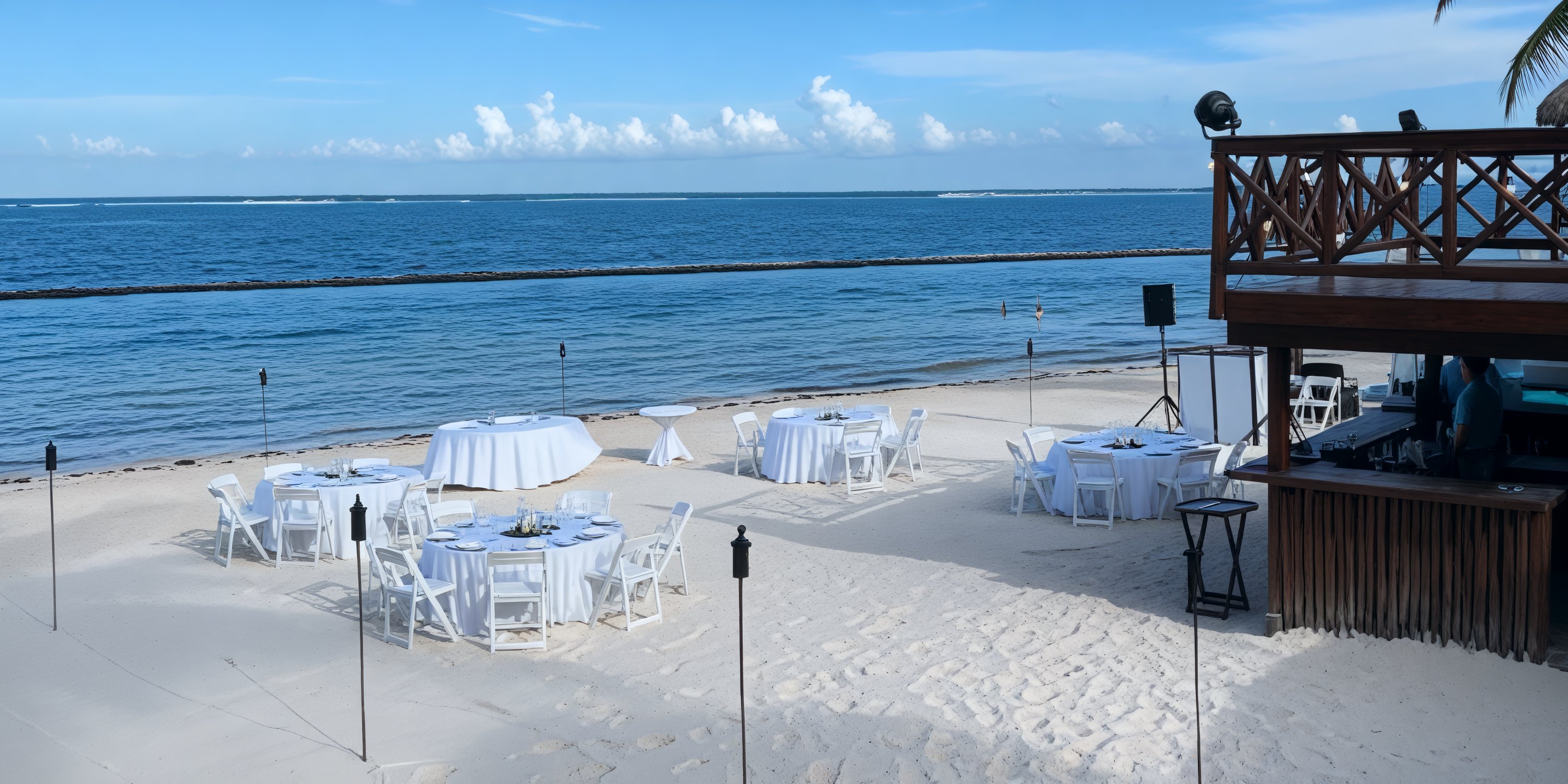 A picturesque destination wedding setup featuring round tables and chairs arranged on the sandy beach by the ocean.